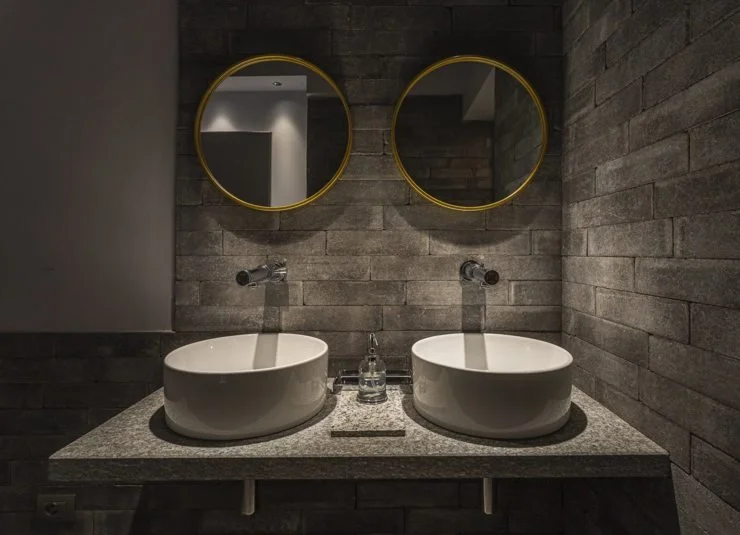 Due lavabi bianchi sul piano in granito con due specchi rotondi sopra, in un bagno con pareti di pietra grigia.
