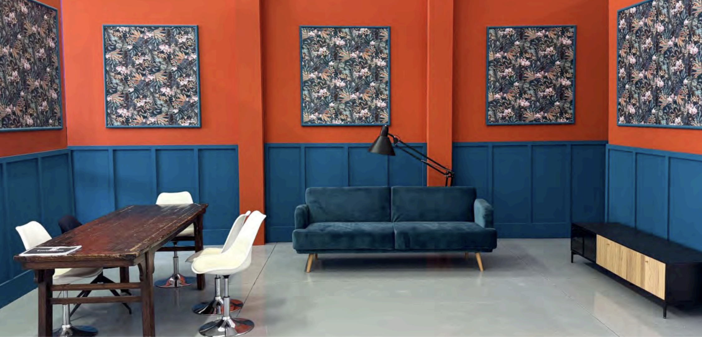 Sala da soggiorno con pareti arancioni e pannelli azzurri, decorata con cinque quadri con motivi floreali, un divano blu-verde, una lampada da terra nera, un tavolo in legno con sedie bianche e nere, e un mobile con ante in legno e struttura nera.