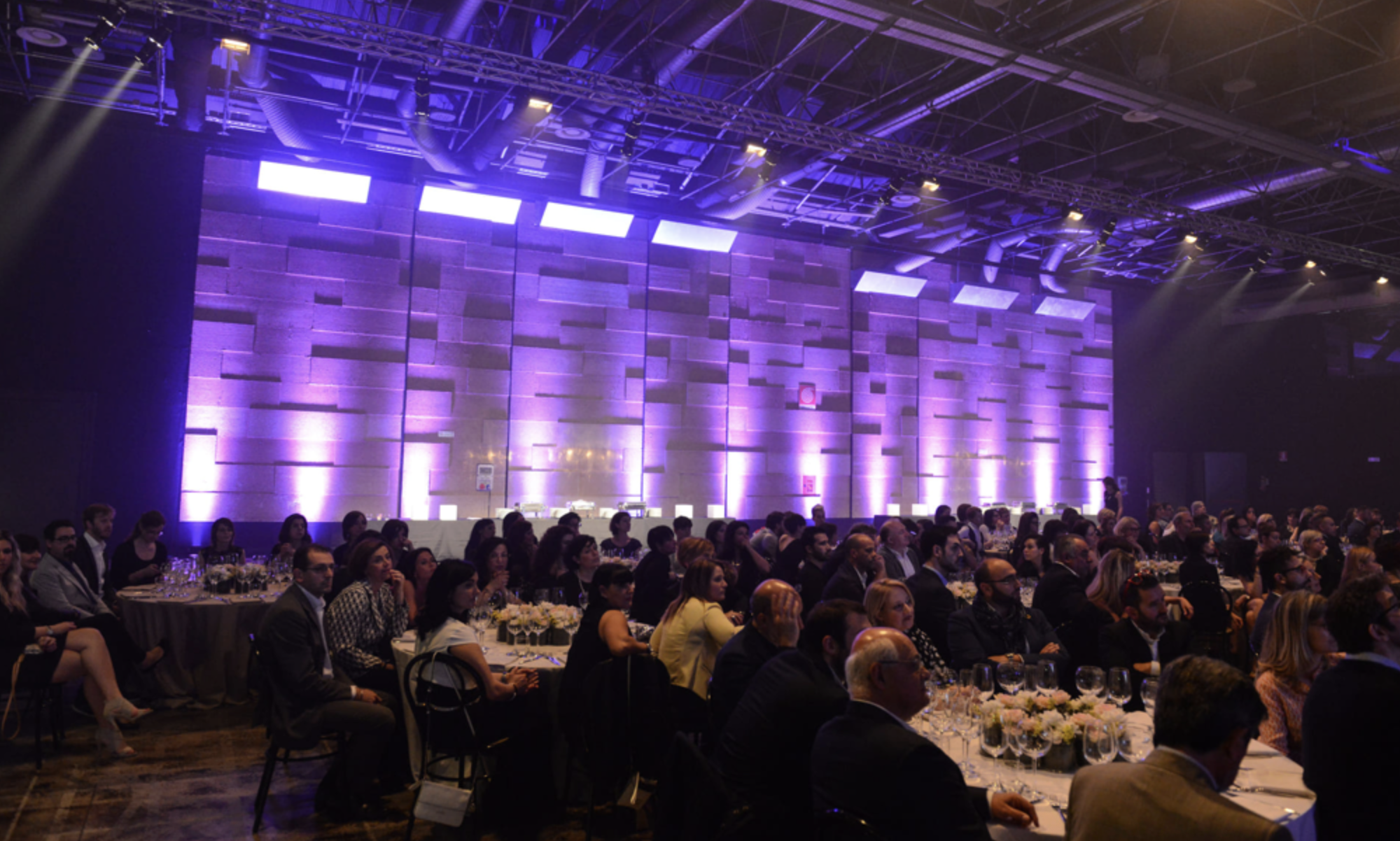 Grande evento in sala con pubblico seduto ai tavoli, illuminazione soffusa e parete di luci viola