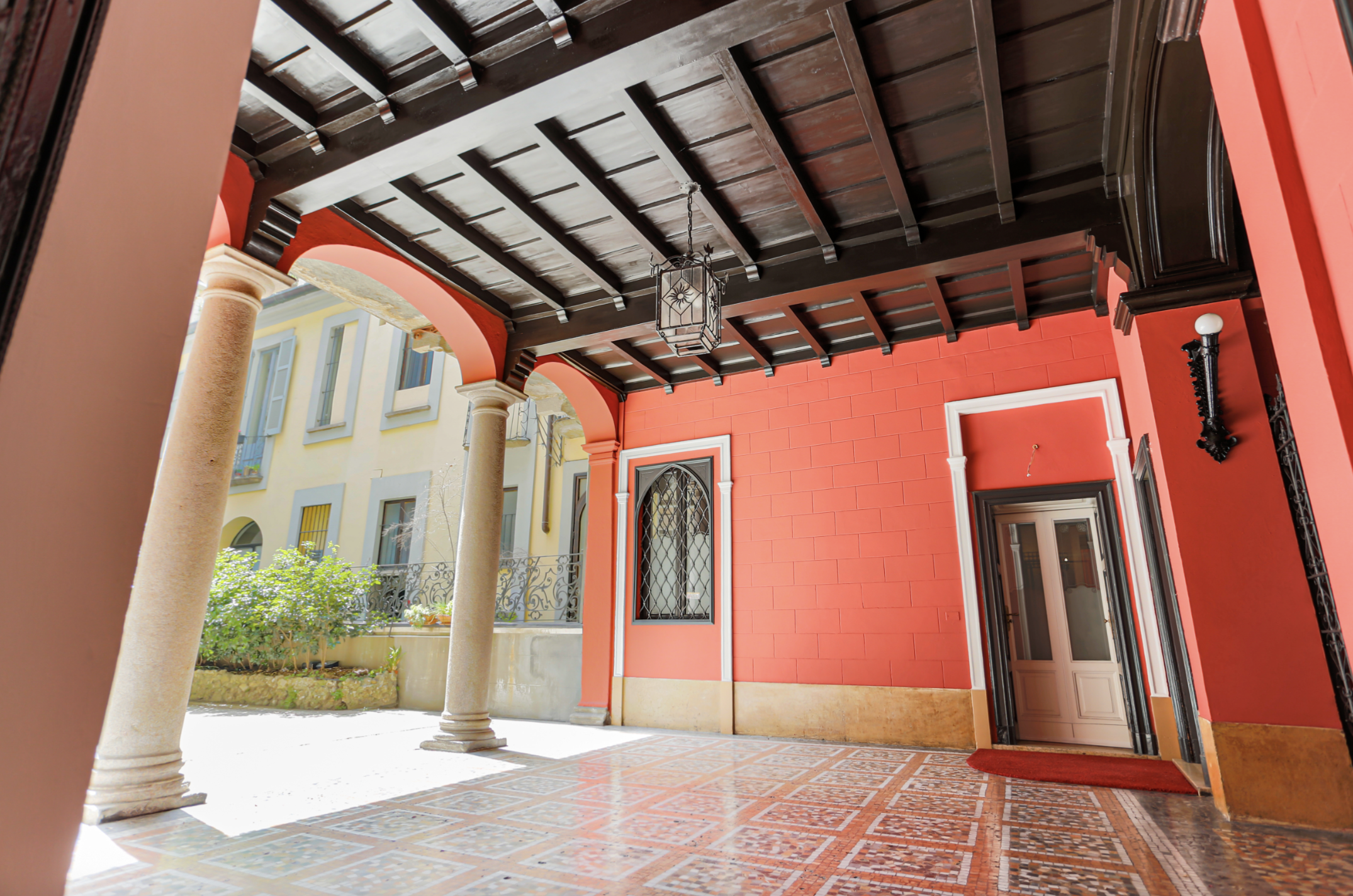 Portico con colonne e soffitto in legno scuro, pareti arancioni e pavimento decorato, con porta e finestra, esposizione di un edificio in più colori.