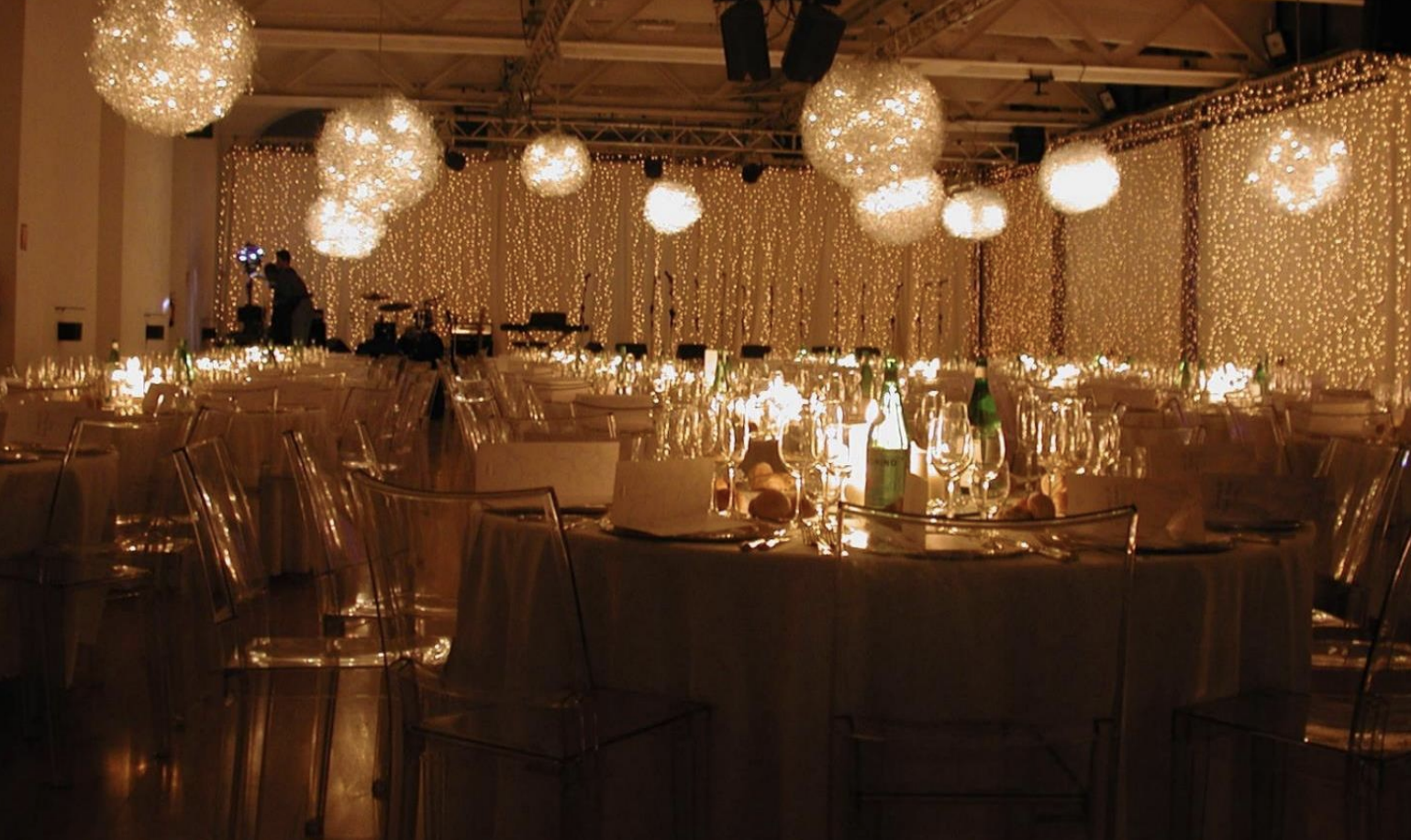 Sala decorata per un evento con luci calde, candelabri, bottiglie di acqua e bicchieri, e sfondo di luci pendenti.
