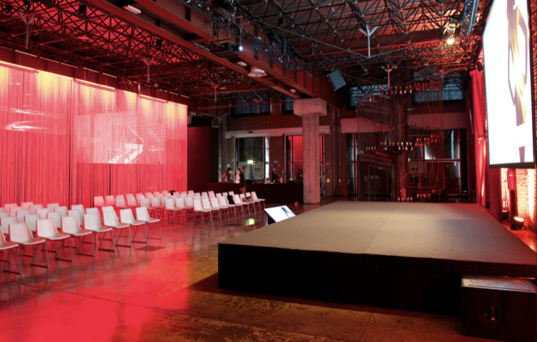 Interno di un locale elegante con luci rosse, una piccola pedana e sedie bianche disposte in fila, pareti con pannelli di tessuto rosso e strutture in acciaio visibili sul soffitto.