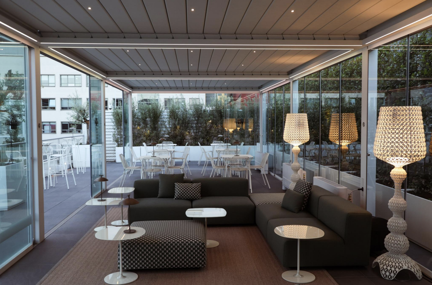 Interno di un bar o sala lounge moderna con divano grigio, lampade decorative luminose, tavolini e una grande finestra che si affaccia su un area esterna con tavoli e sedie