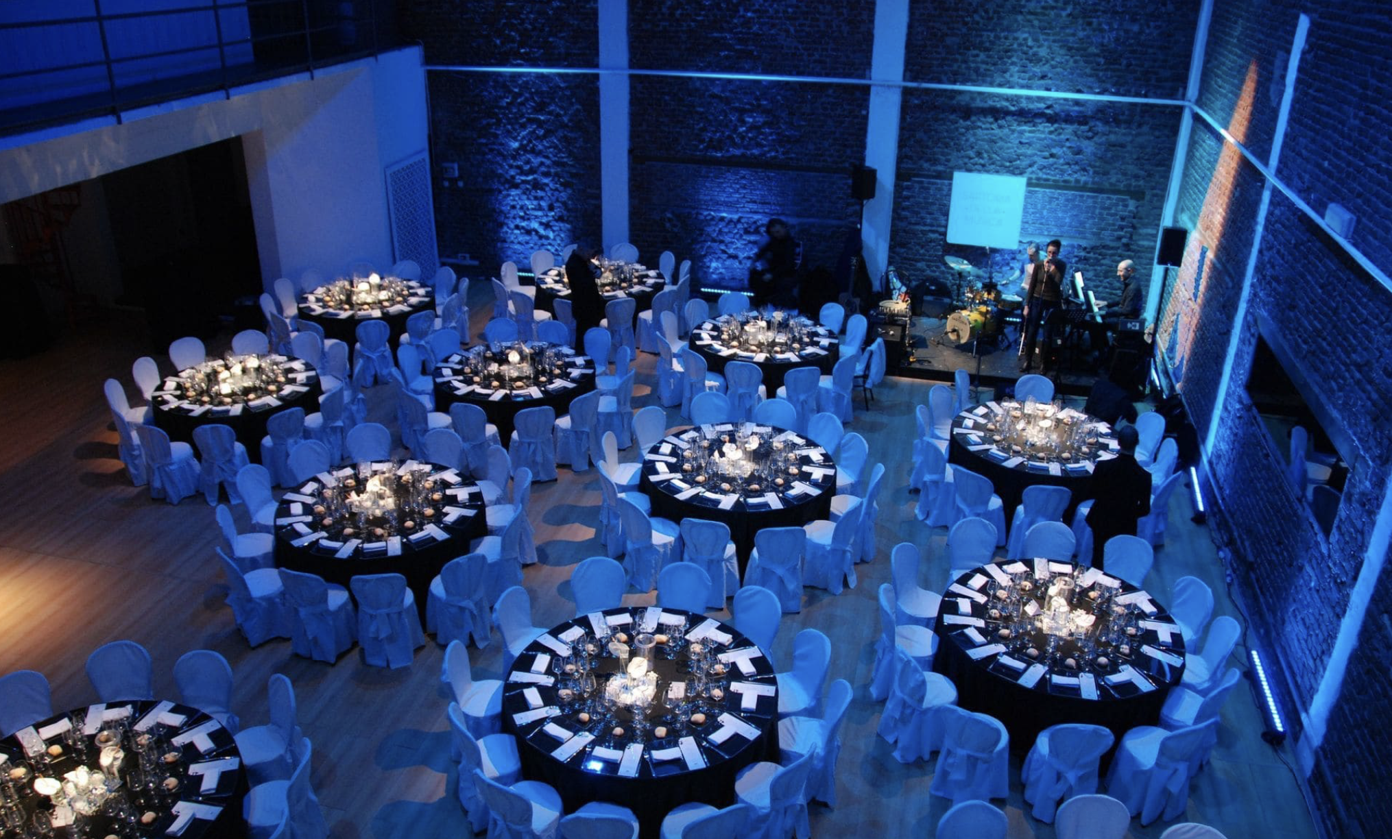 Sala di eventi elegante con tavoli rotondi coperti da tovaglie nere, decorati con candele e centrotavola, illuminata da luci blu, con personnel e band musicale sul palco.
