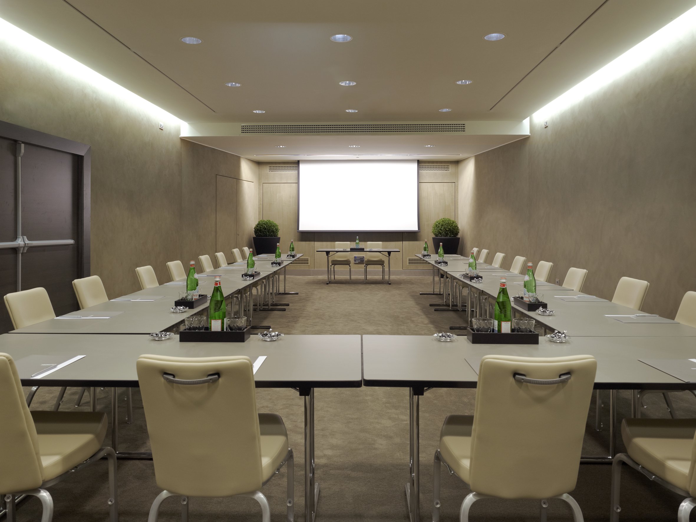 Sala riunioni con tavoli a forma di U, sedie beige, bottiglie d'acqua, bicchieri e blocchi note, grande schermo bianco sul muro opposto e piante decorative ai lati.