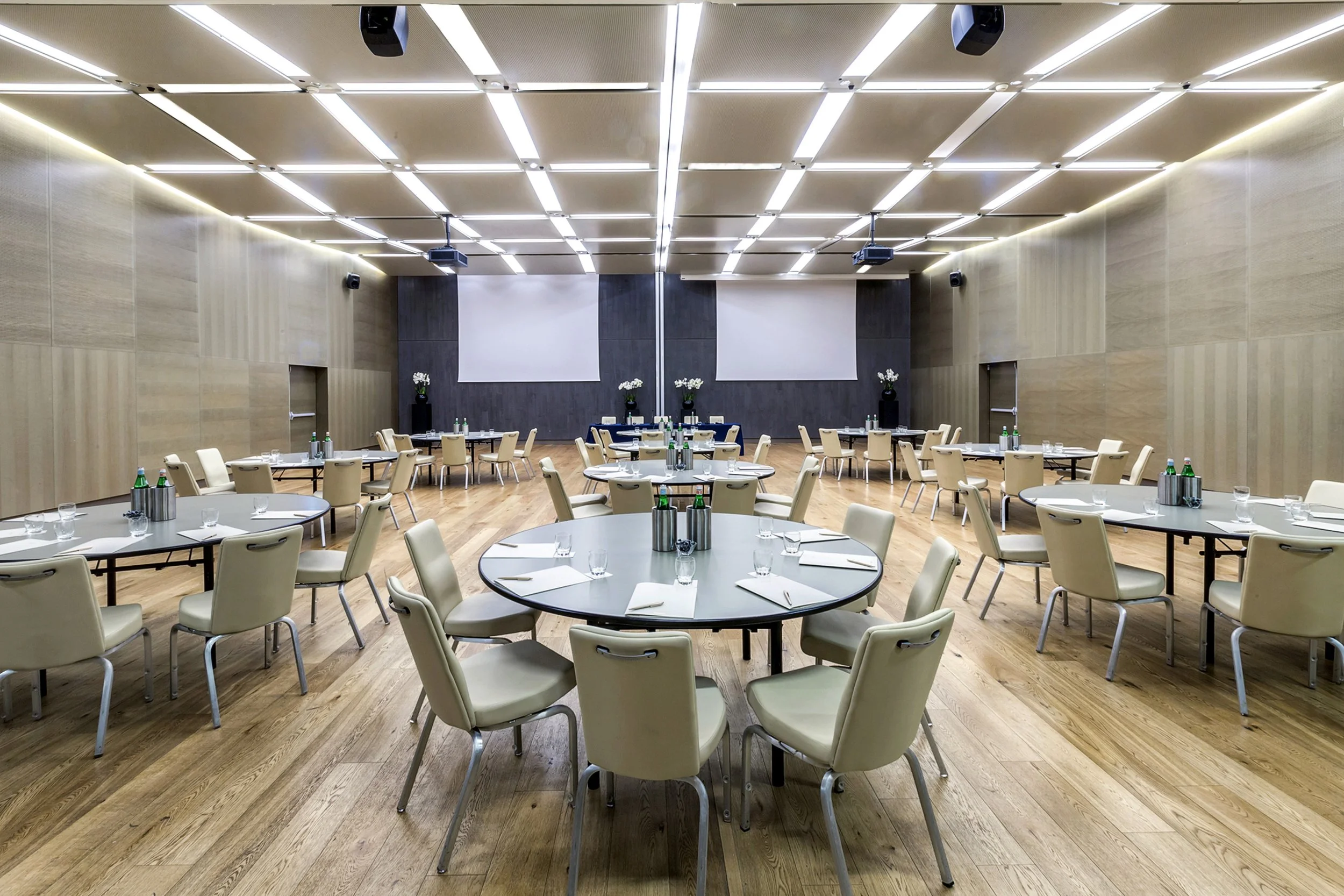 Sala per conferenze con tavoli rotondi e sedie chiare, decorata con vasi di fiori bianchi e bottiglie d'acqua minerale, con schermi e proiettori sul soffitto.