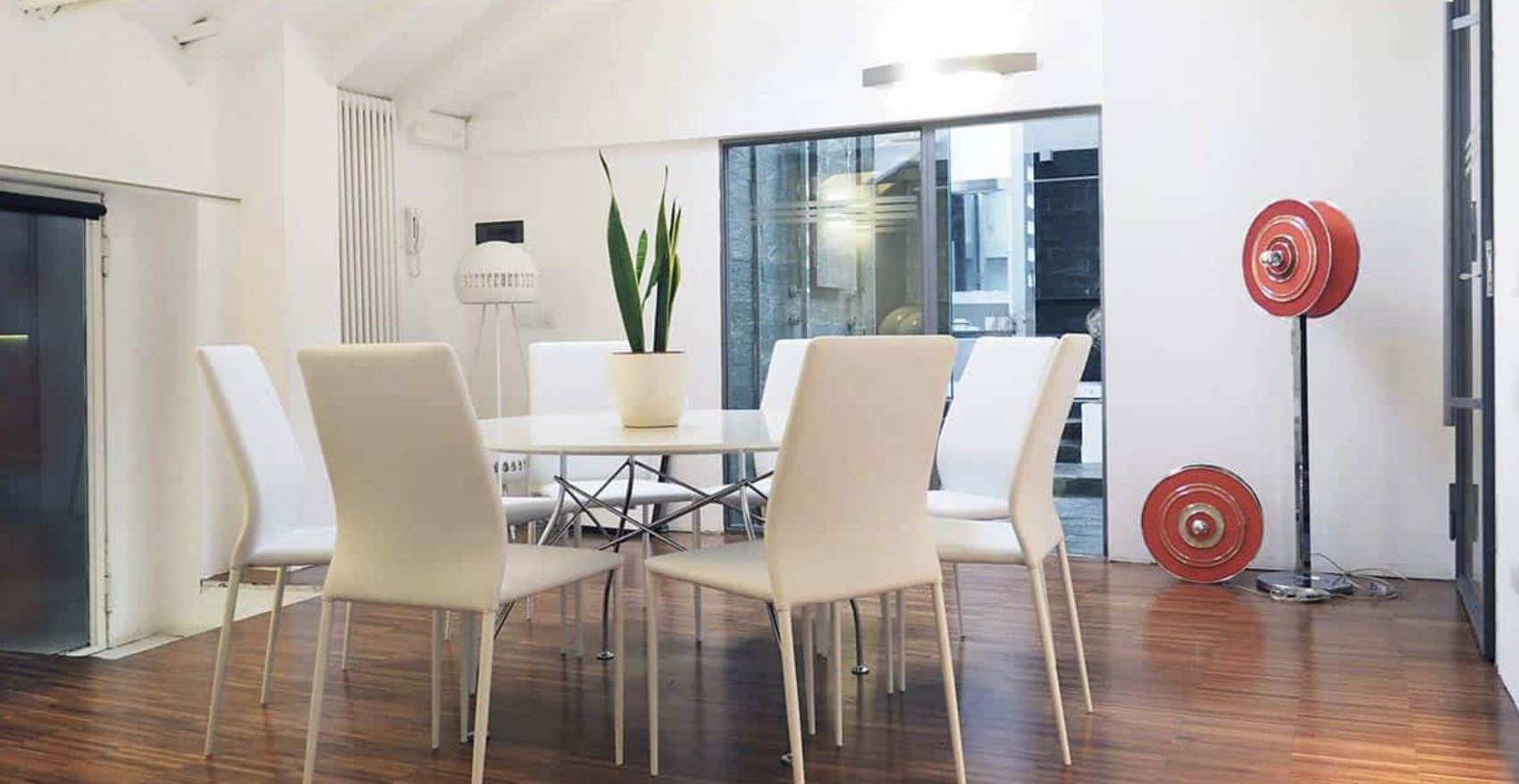 Sala da pranzo moderna con tavolo rotondo, sei sedie bianche, vaso con pianta al centro, lampada da terra, due pesetti rossi e una grande finestra con vista a un cortile esterno.