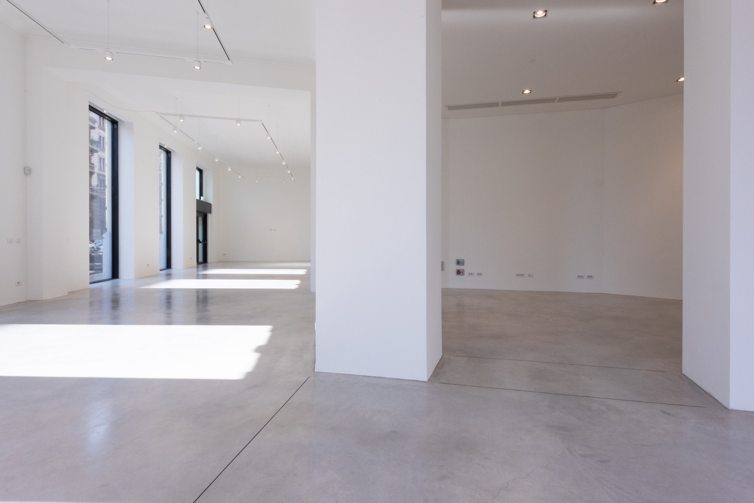 Interno vuoto di un edificio con pareti e soffitto bianchi, grandi finestre con luce naturale e pavimento in cemento, pareti a muro diviso che crea due ambienti distinti.