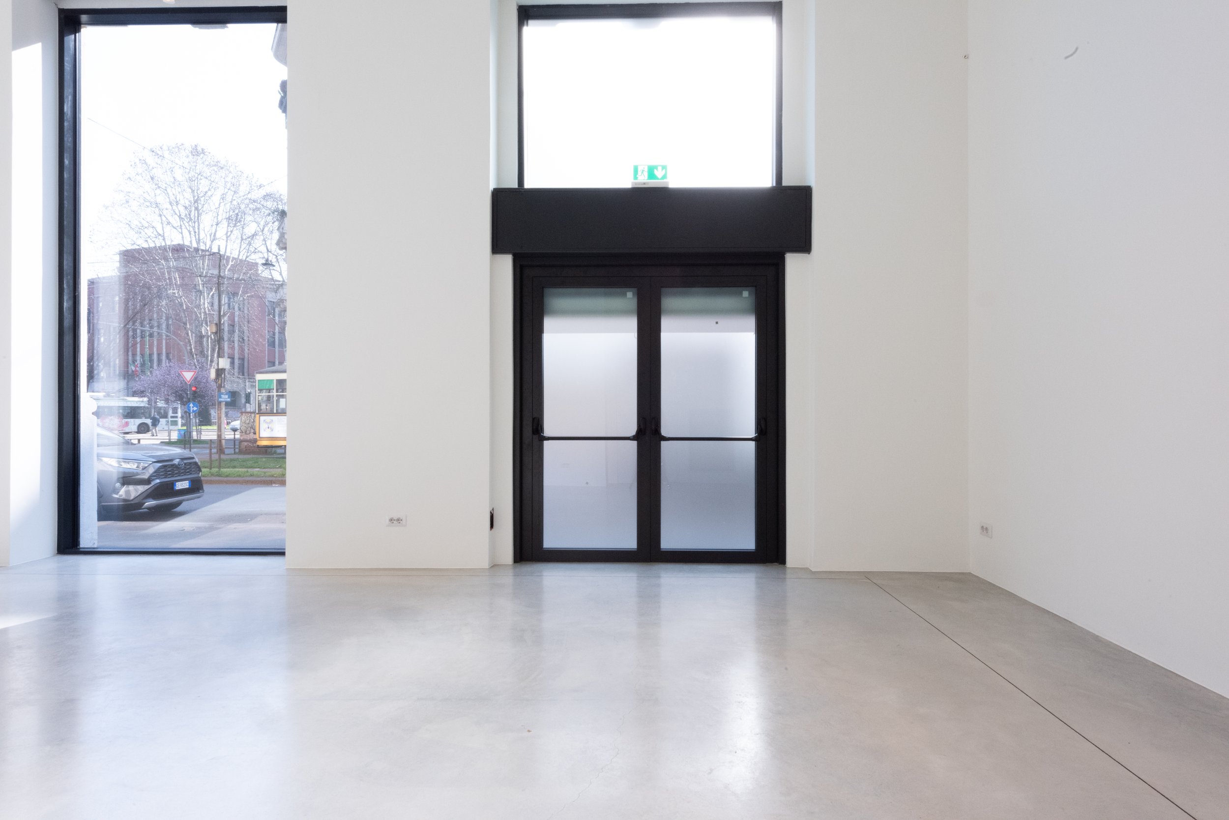 Interno di un edificio moderno con pareti bianche e pavimento in cemento, grande porta scorrevole e finestra con vista sulla strada esterna.