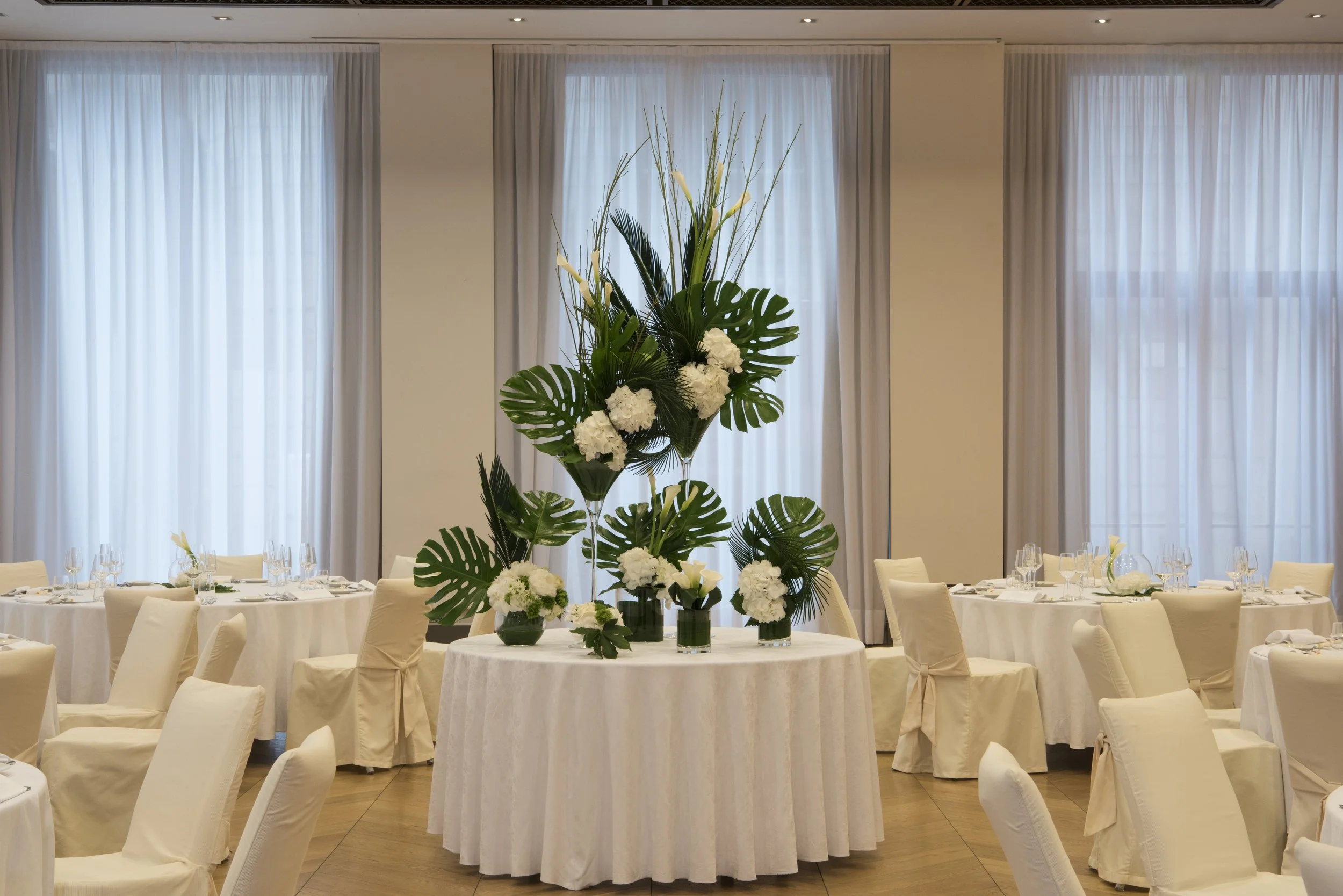 Decorazione floreale con grandi foglie di monstera e fiori bianchi su un tavolo rotondo in una sala elegante con tende bianche e tavoli apparecchiati con tovaglioli e bicchieri.