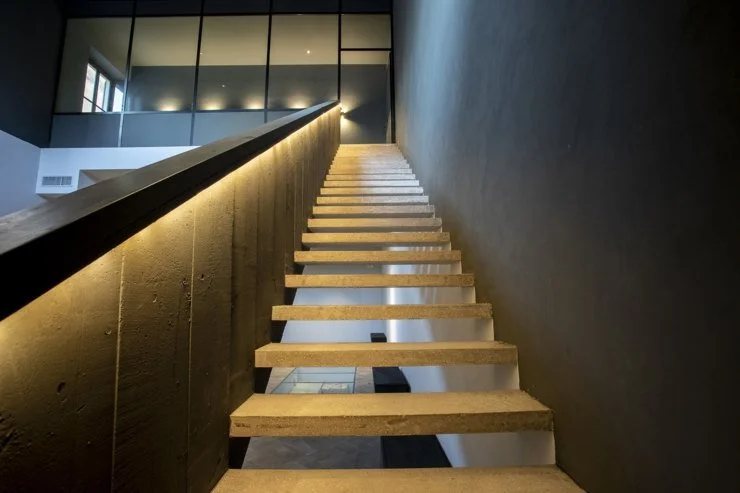 Scala moderna in legno con luce integrata, interno di un edificio con pareti grigie e grandi finestre.