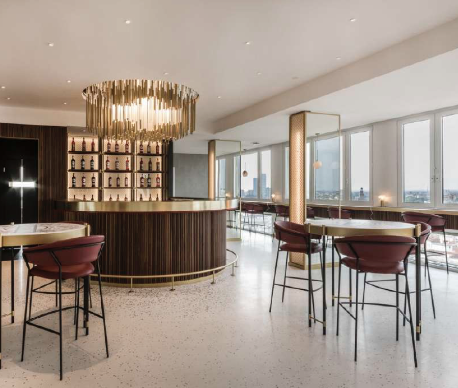 Interno di un bar o ristorante elegante con luci soffuse, bancone in legno scuro, scaffali con bottiglie alle spalle, tavoli rotondi con sedie di velluto rosso e grandi finestre che mostrano una vista urbana.