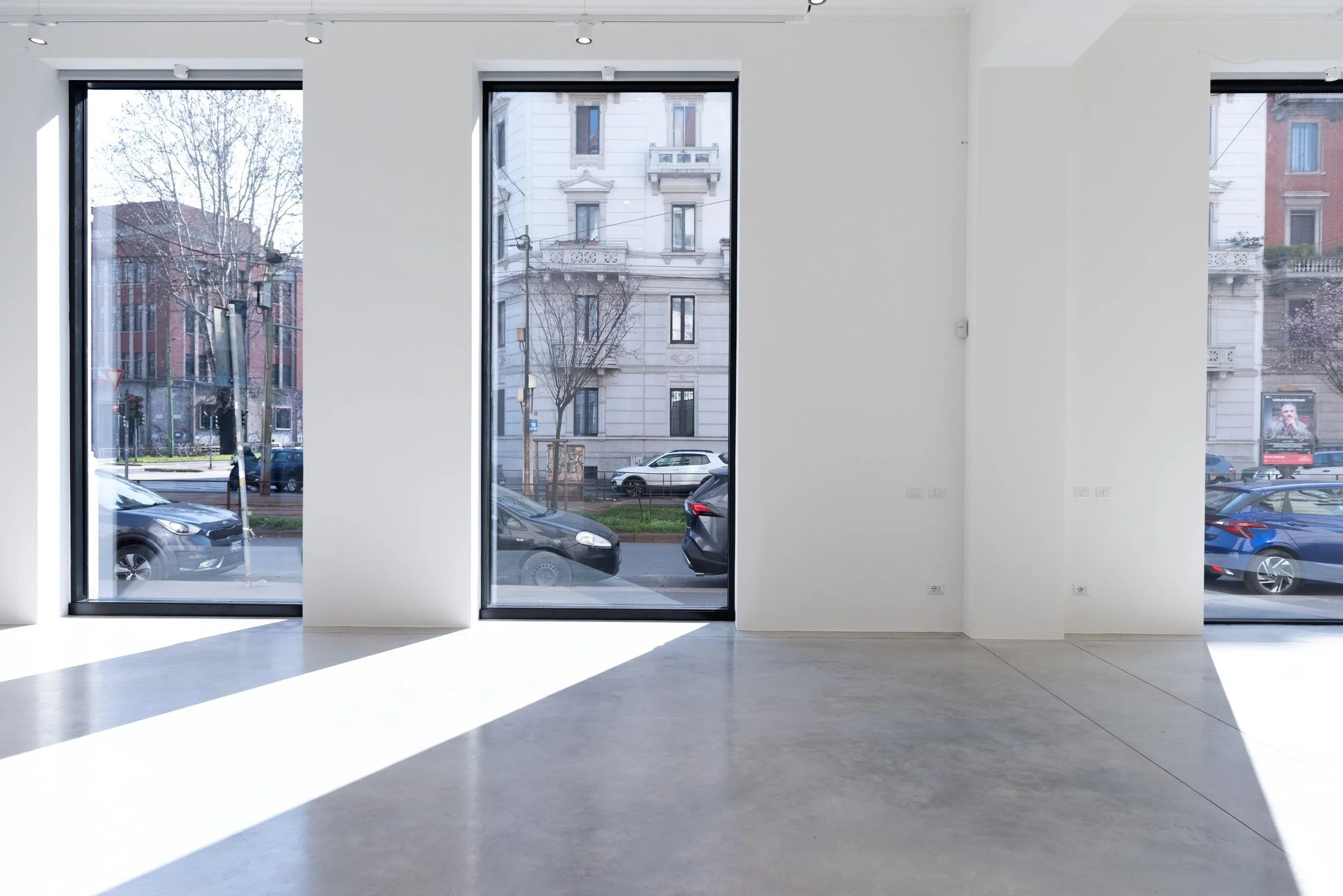 Interno di una stanza vuota con grandi finestre che danno sulla strada. All'esterno si vedono macchine parcheggiate e edifici con balconi e finestre.