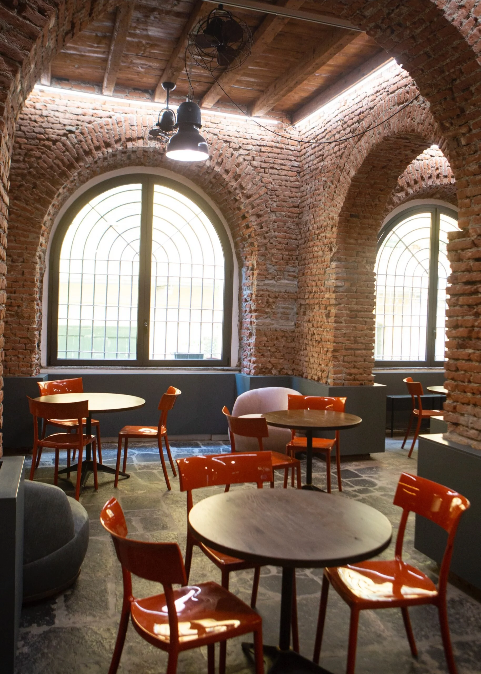 Interno di un caffè con pareti in mattoni rossi, archi in mattoni, grandi finestre ad arco con grate, illuminazione soffusa e sedie arancioni attorno a tavoli di legno.