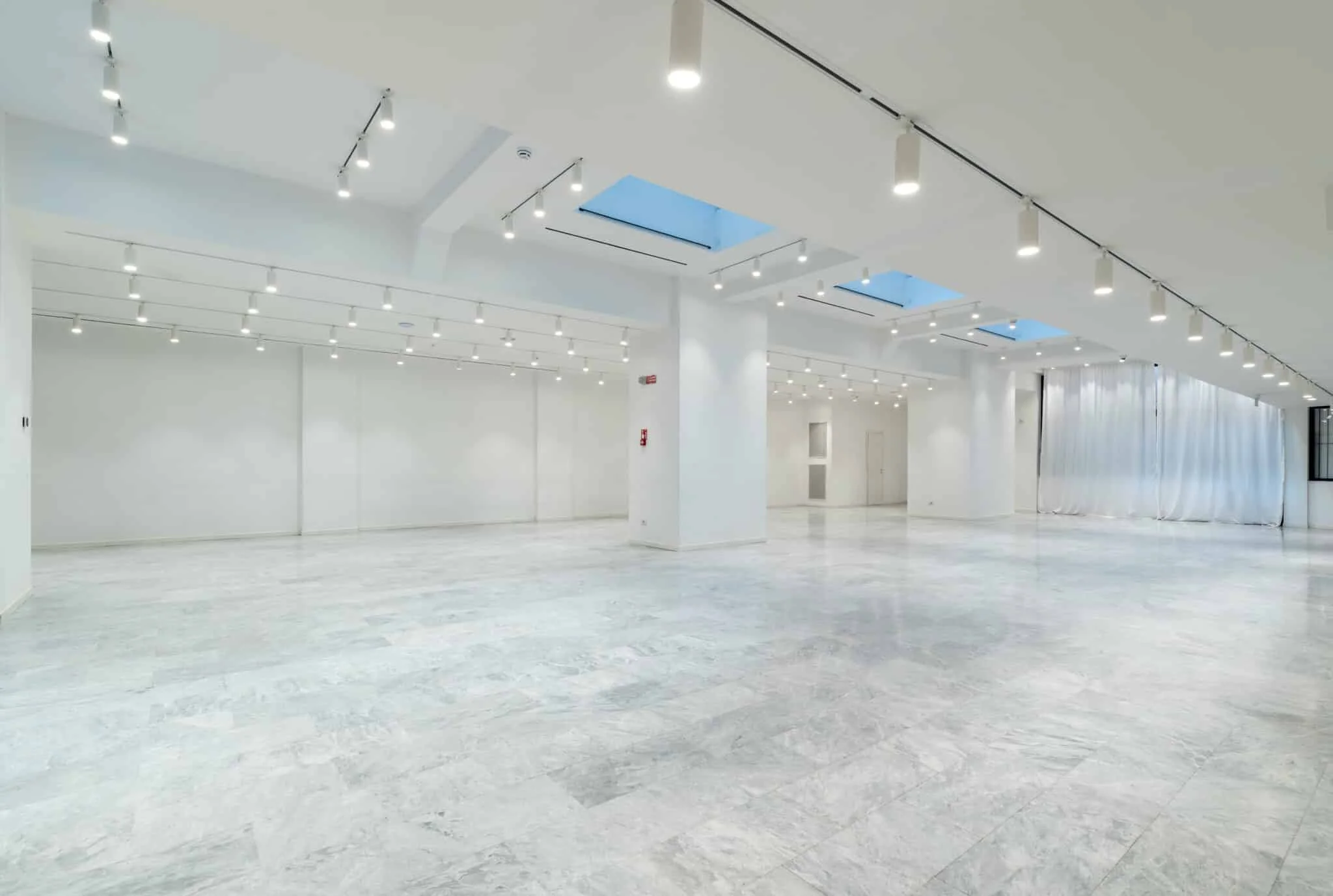 Grande spazio vuoto con pareti bianche, pavimento in marmo chiaro, luci al soffitto, grandi finestroni con tende bianche.