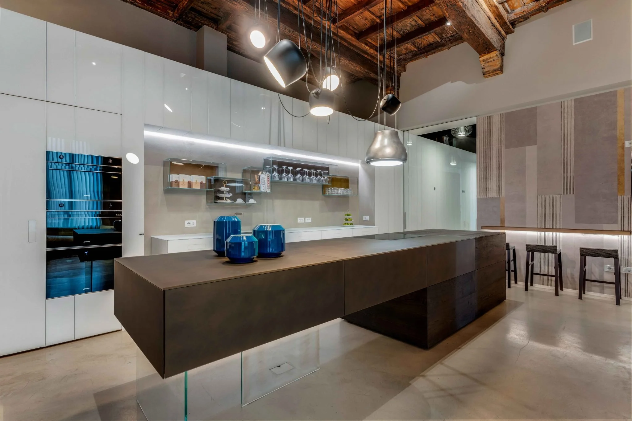 Cucina moderna con isola di legno scuro, lucida e decorata con cinque vasi blu. Pareti biane con armadietti a muro, alcune tazzine e bicchieri su mensole di vetro. Illuminazione sospesa sopra l'isola, soffitto in legno a vista.