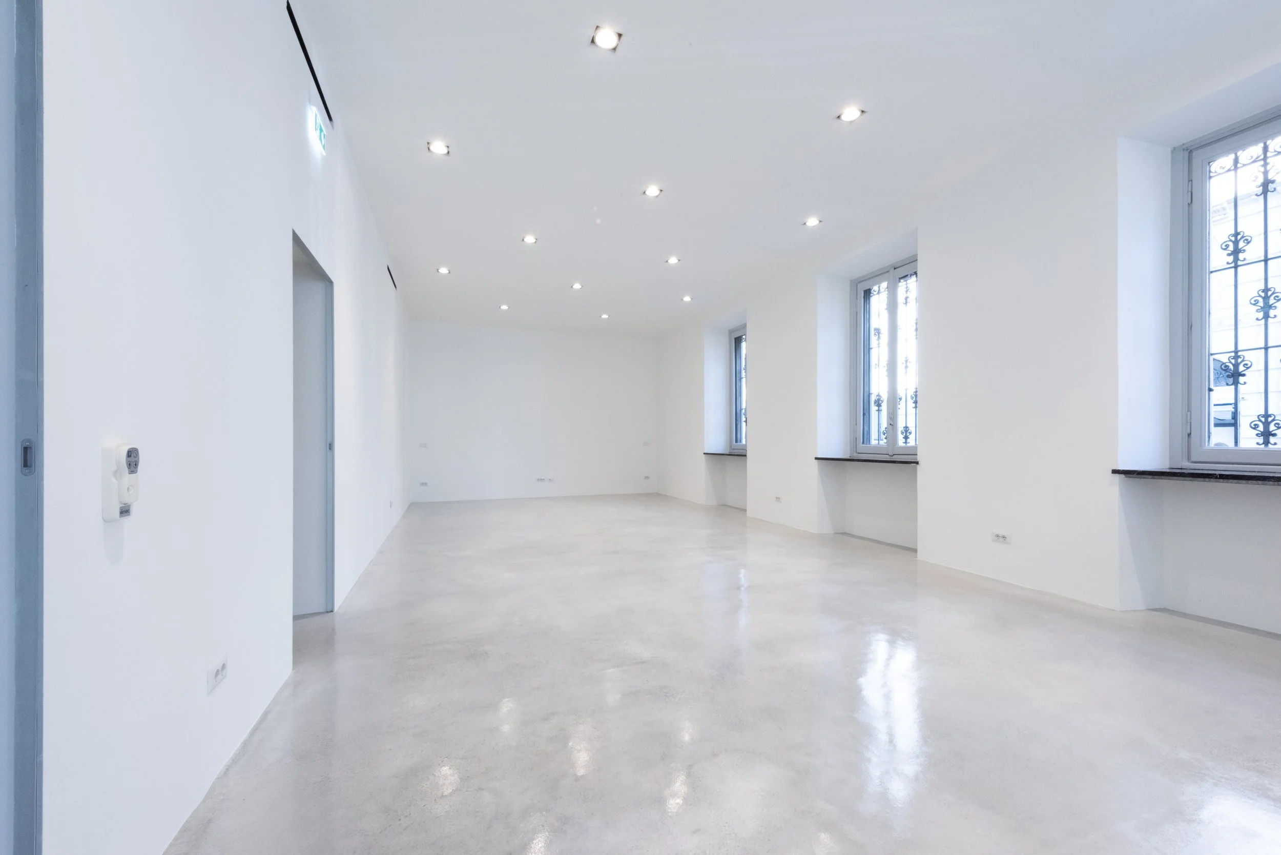 Interno di una stanza spaziosa con pareti bianche, pavimento in cemento lucido, quattro grandi finestre con inferriate e luci del soffitto.