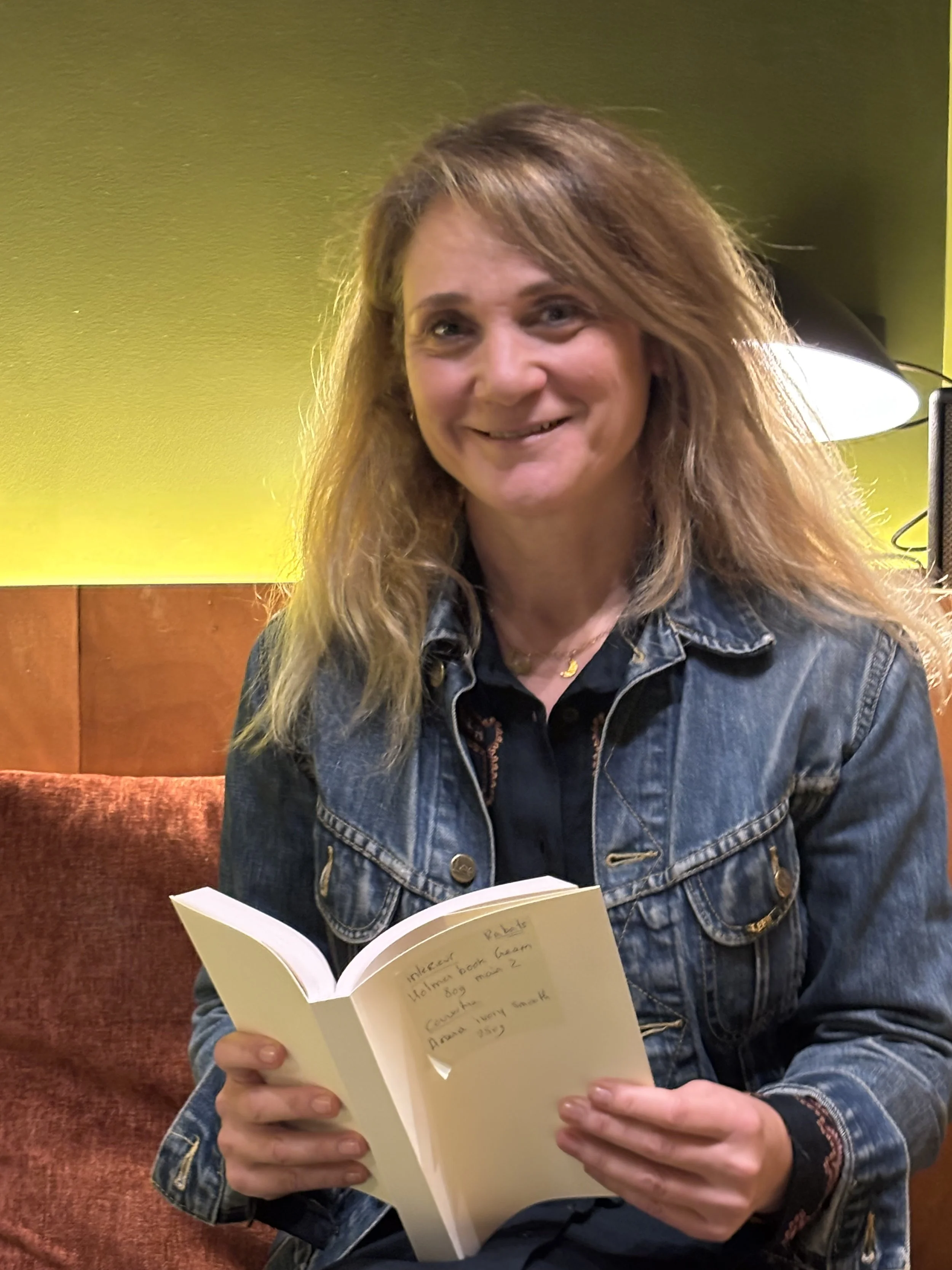Une femme souriante portant une veste en jean, assise sur un canapé, tenant un livre avec une note à l'intérieur.