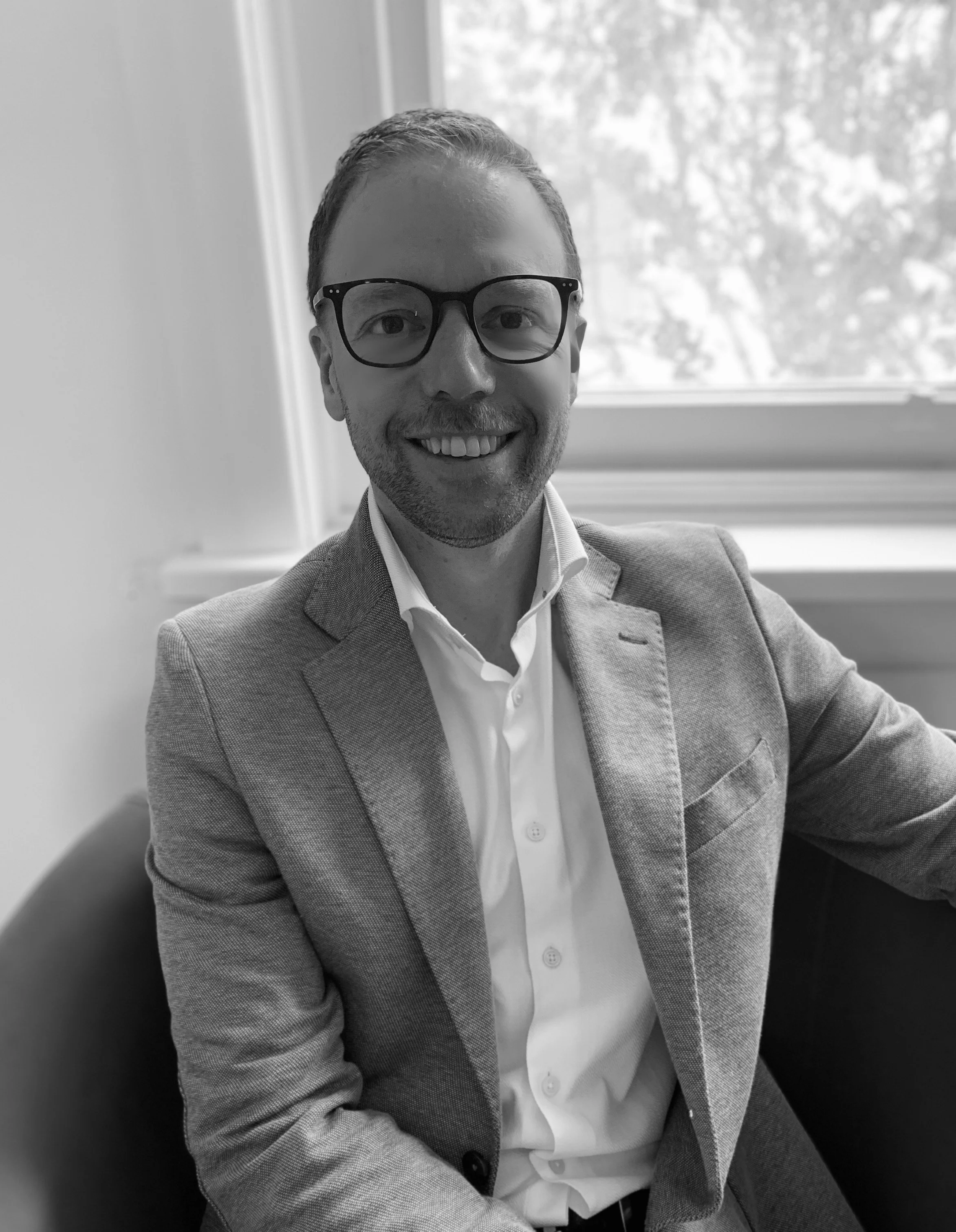 Black-and-white headshot of Dr Dominic Doyle, Clinical Psychologist, in his office, smiling at the camera, wearing glasses, a jacket and shirt