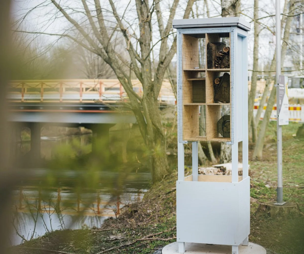 A tall, light blue wooden insect hotel with various nesting materials standing near a riverbank with a bridge in the background.