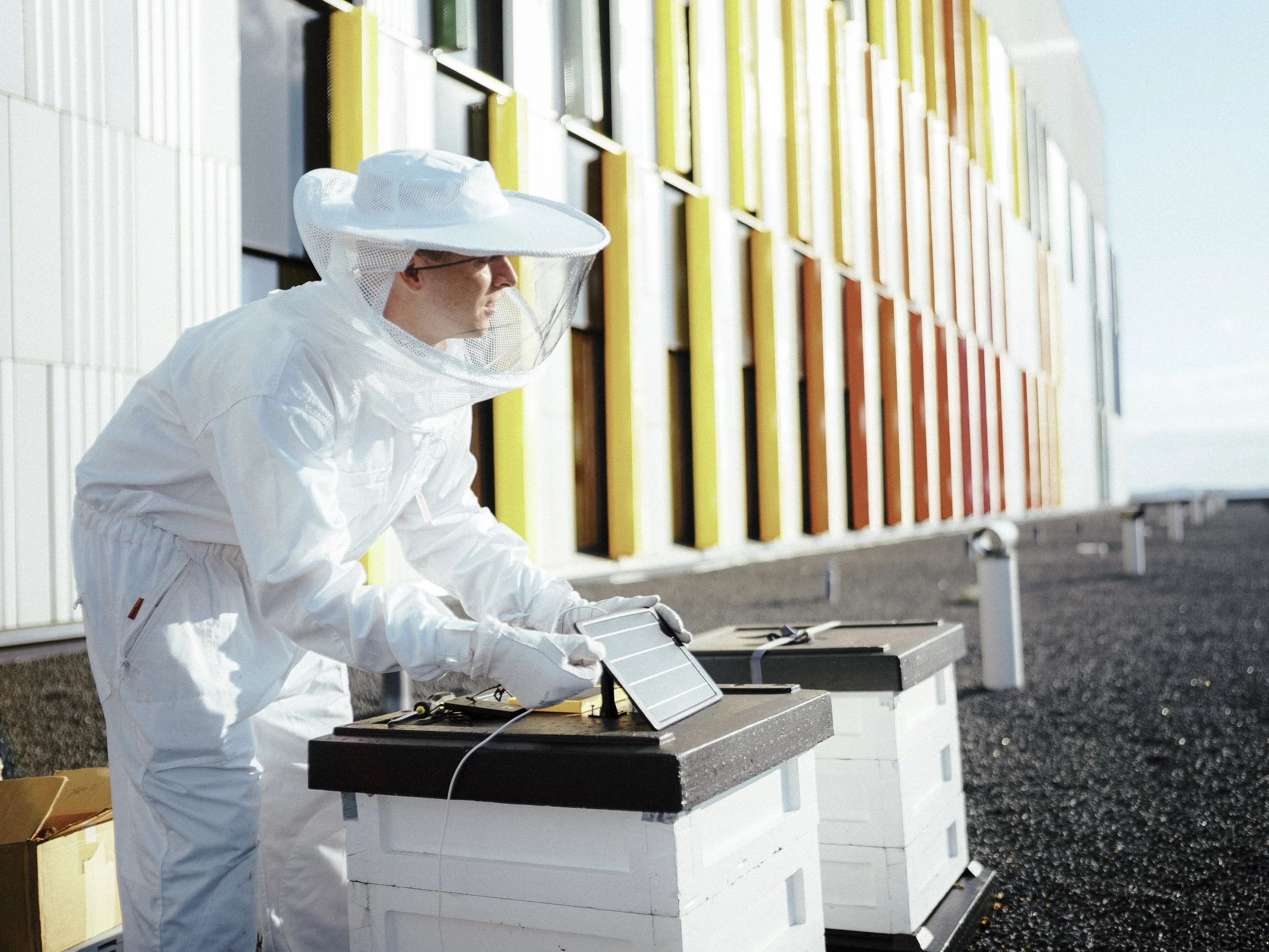 A person in a white beekeeping suit working on a beehive equipped with a small solar panel on a modern rooftop.