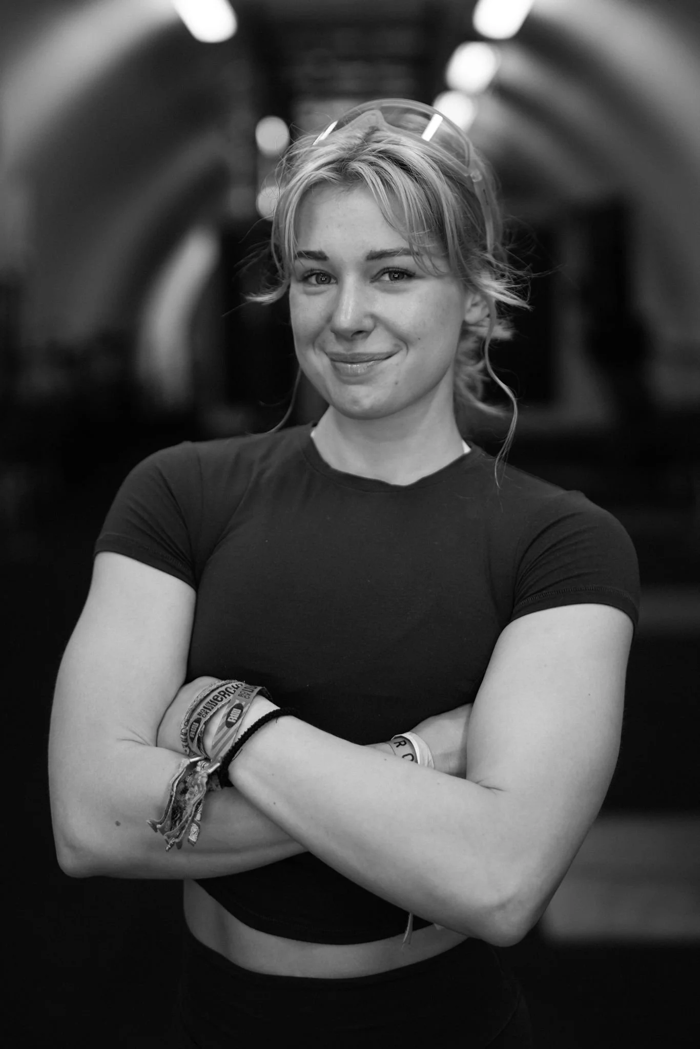 Black and white photo of a young woman with short hair, smiling, with arms crossed, wearing a black t-shirt and wristbands, standing in an indoor tunnel or corridor.
