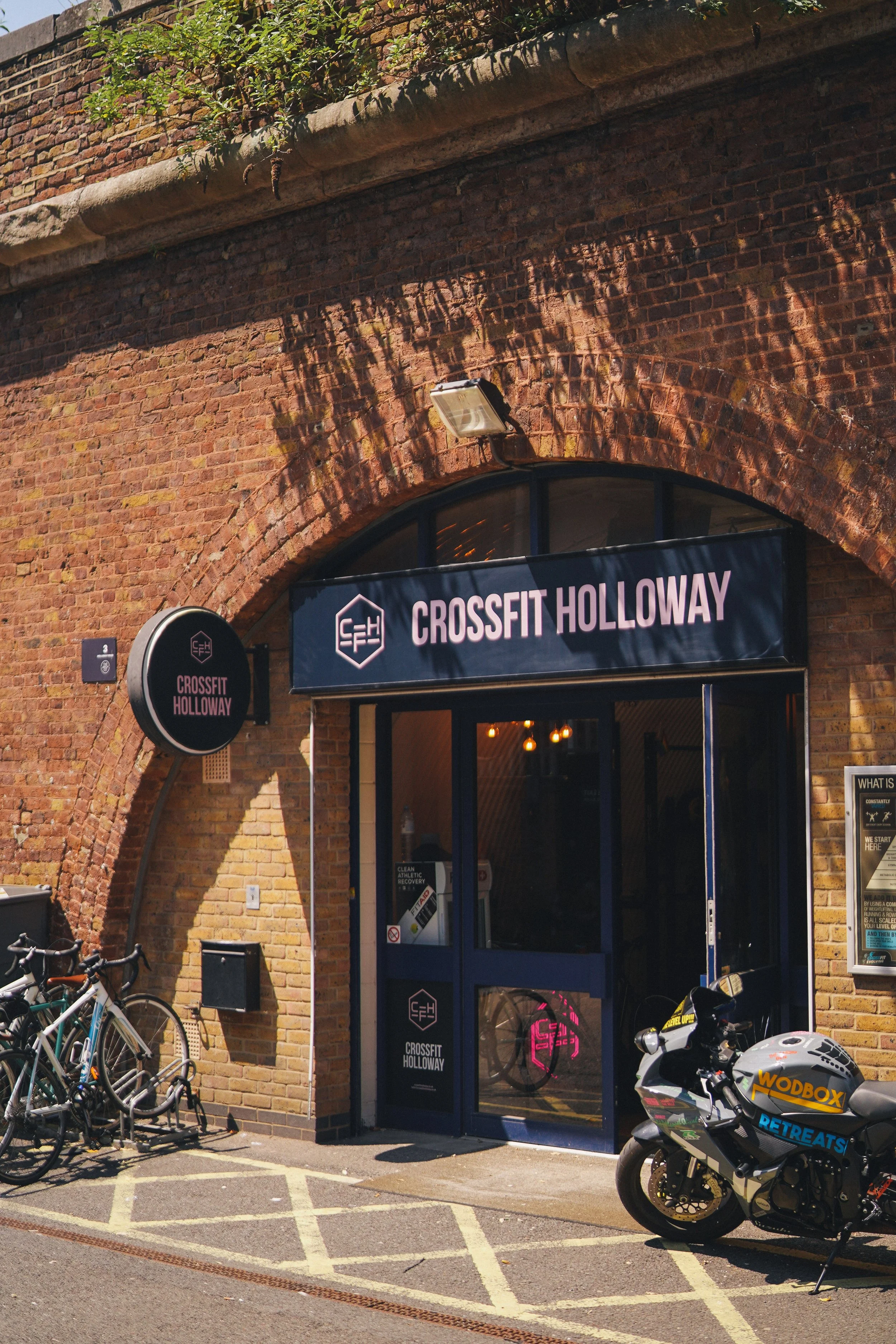 Entrance of Crossfit Holloway gym in a brick building with bikes parked outside and a motorcycle nearby.