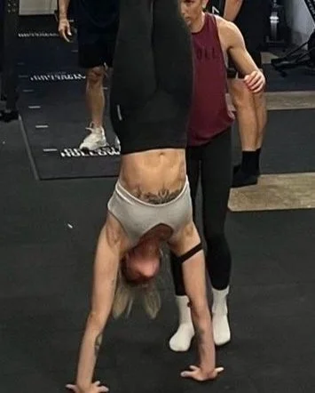 Person performing a handstand in a gym with others around, one person watching.