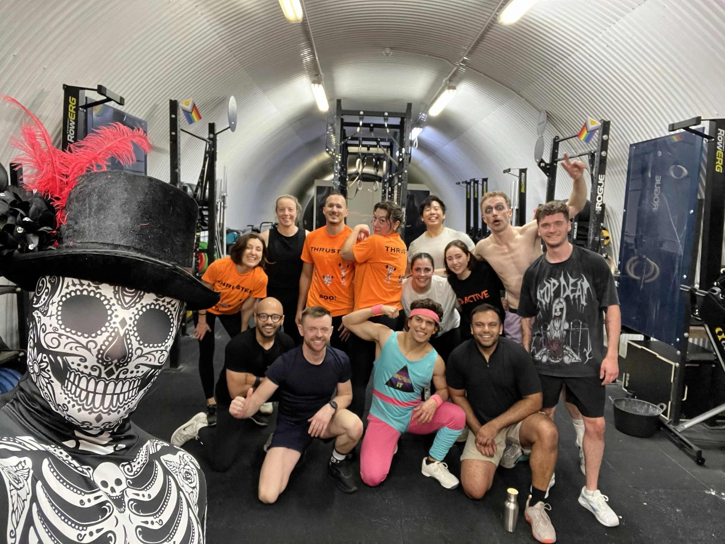 Group of people at a gym, smiling and posing after a workout, with a skeleton figure wearing a black top hat with red feathers in the foreground.