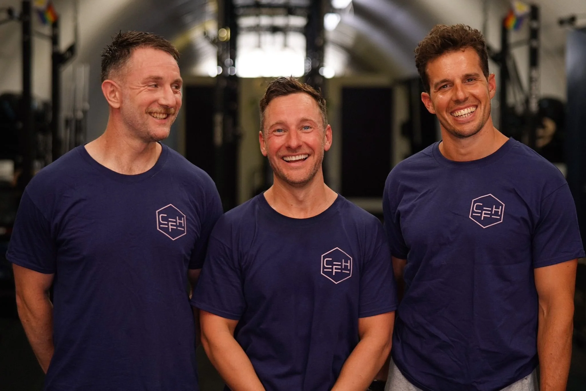Three men in matching navy blue t-shirts with a white logo, smiling and standing side by side in a gym or fitness center.