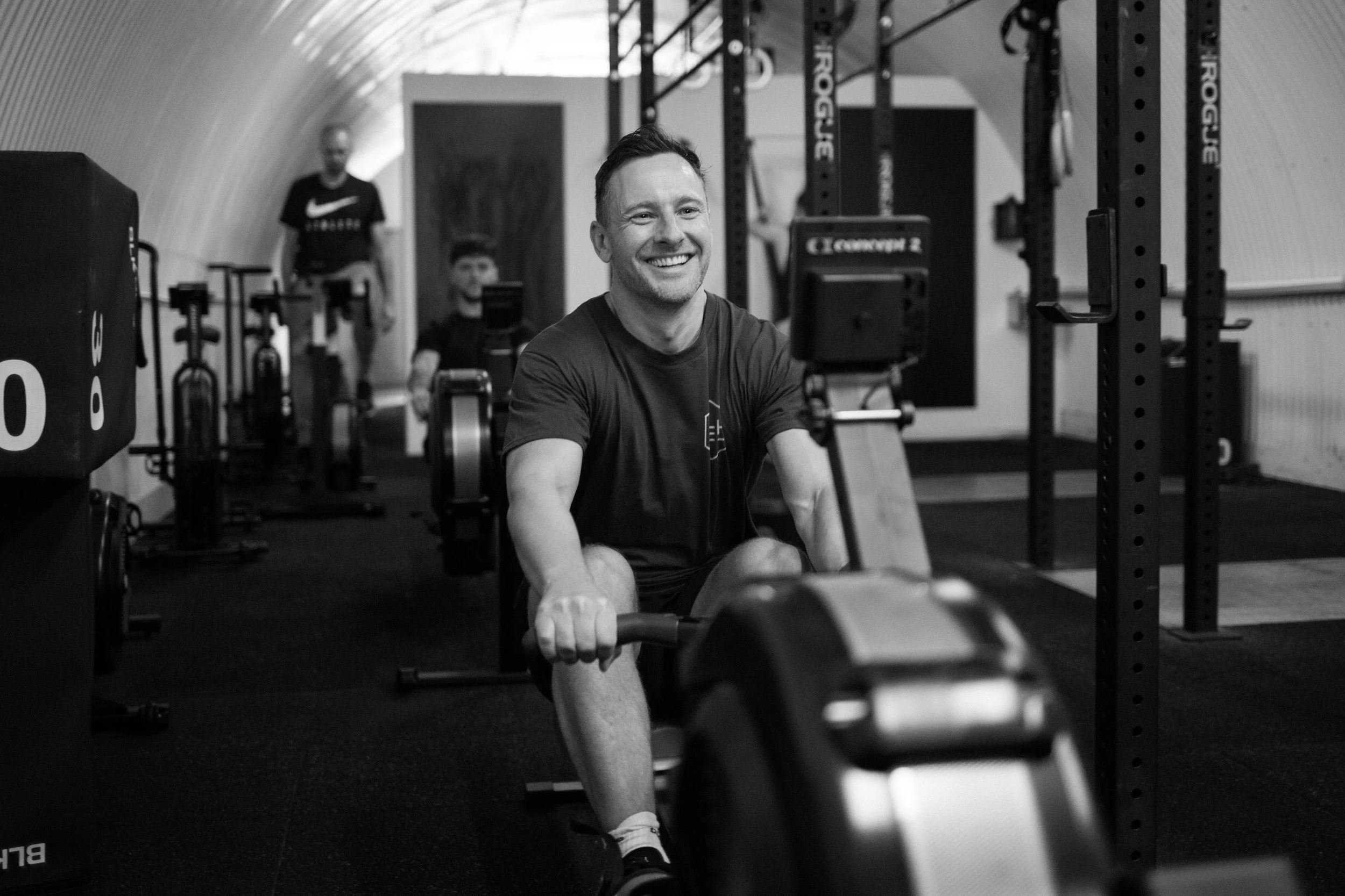 A man smiling while rowing a machine in a gym with other people in the background.