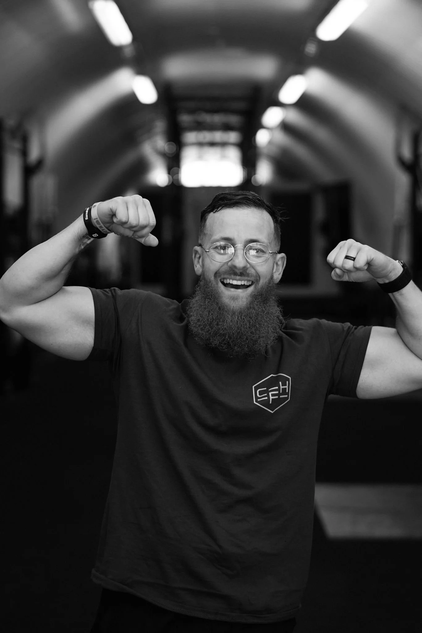 A smiling man with a beard and glasses flexing his biceps in a tunnel with a high, arched ceiling.