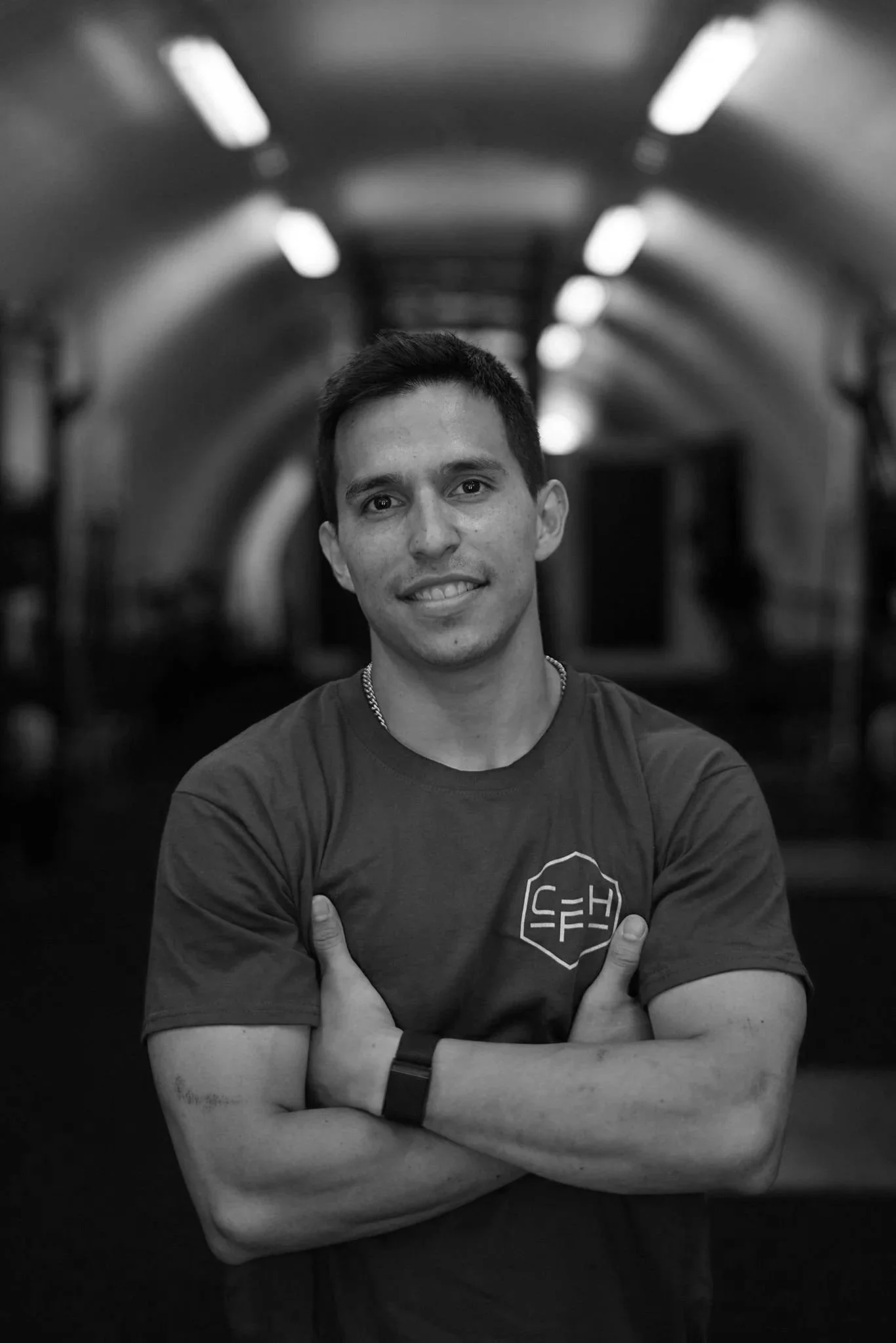A young man with short dark hair and a smile, crossing his arms, wearing a t-shirt with a logo, in an indoor tunnel with lights overhead.