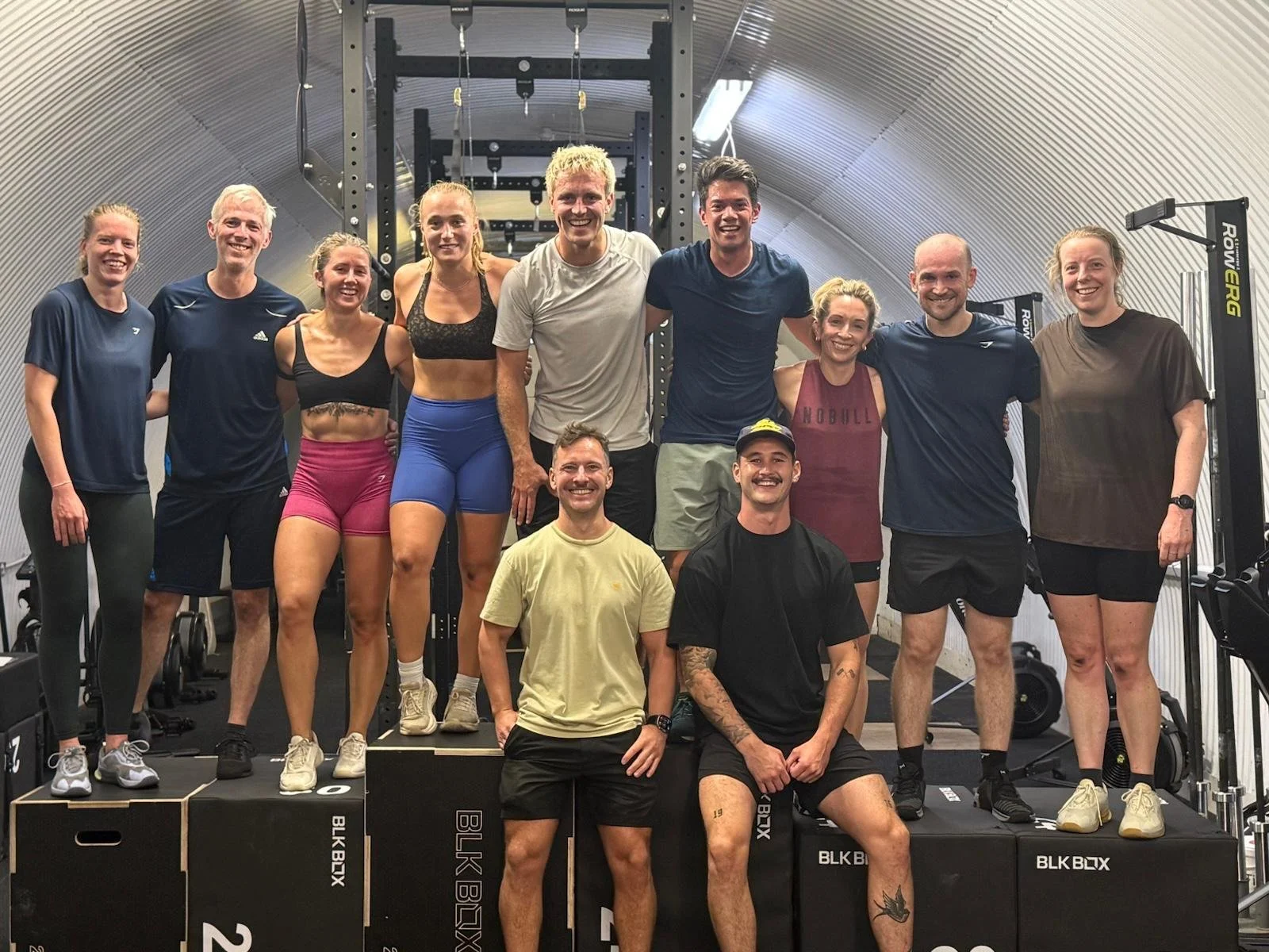Group of ten people in a gym posing together, some standing on workout platform boxes, smiling at the camera.
