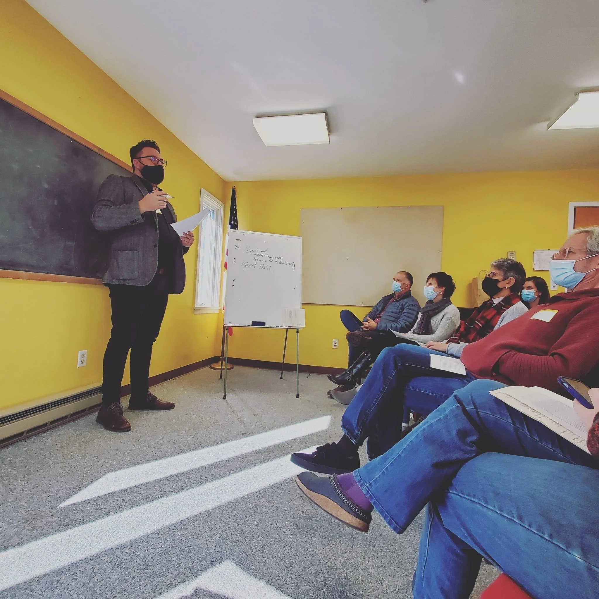 A man in a grey blazer and black mask giving a presentation in a classroom, with several seated people wearing masks listening to him.
