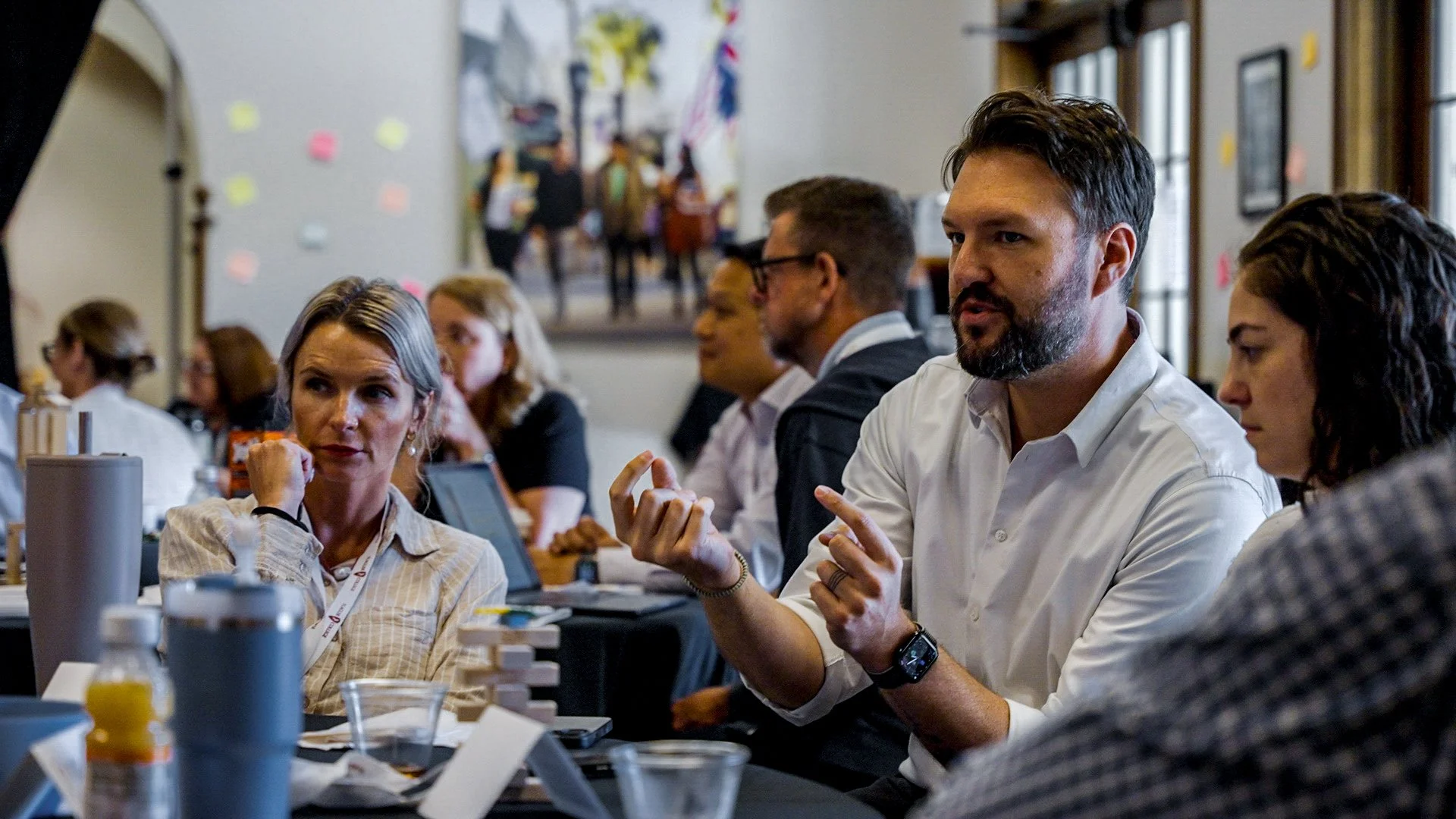 People attending a conference or seminar, sitting around a table in a well-lit room with large windows, engaged in discussion.