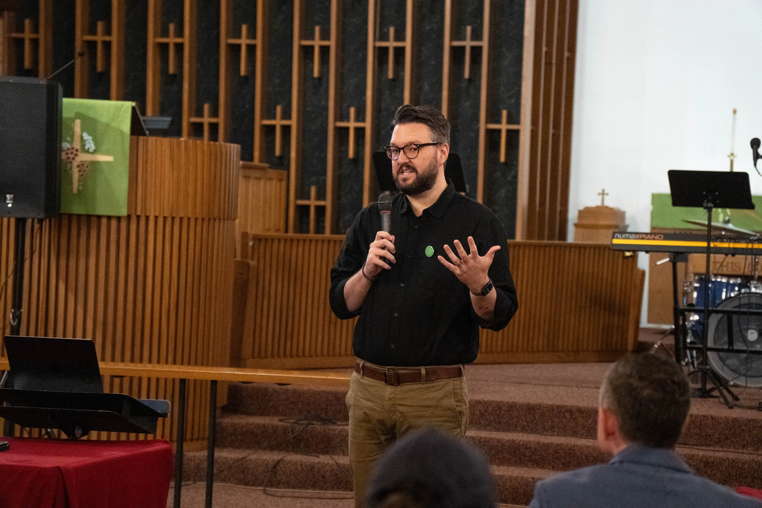 A man with glasses, a beard, and dark hair, wearing a black shirt and khaki pants, is holding a microphone and speaking to an audience in a church or auditorium setting with wooden decor and musical instruments in the background.
