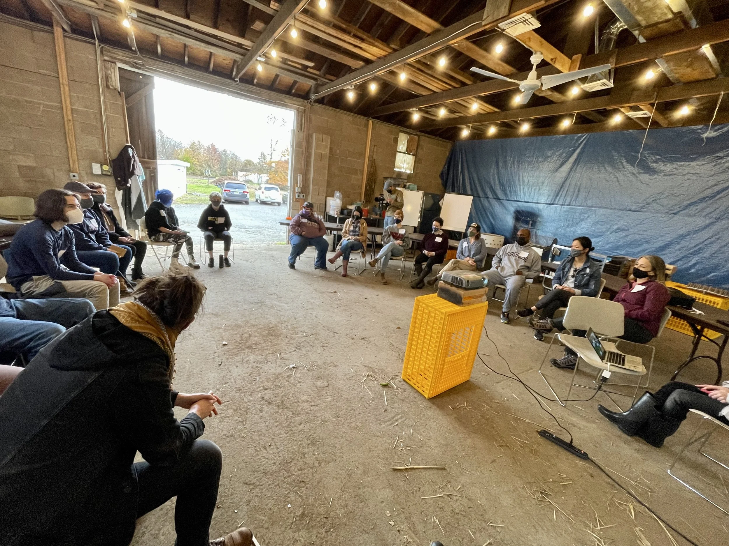 A group of people is seated in a semi-circle inside a barn, attending a meeting or workshop, with some wearing masks. The barn has a high wooden ceiling with string lights, and the open doors reveal a parking lot and outdoor scenery.