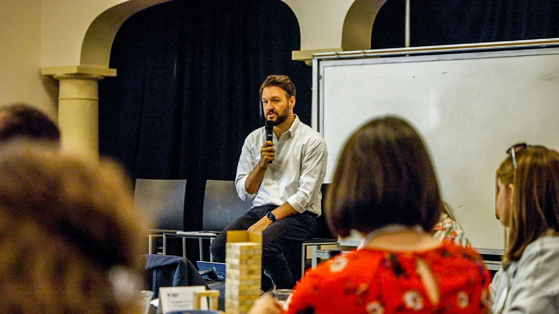 A man with a beard and white shirt sits on a table holding a microphone, speaking to an audience in a room with dark curtains and a whiteboard in the background.