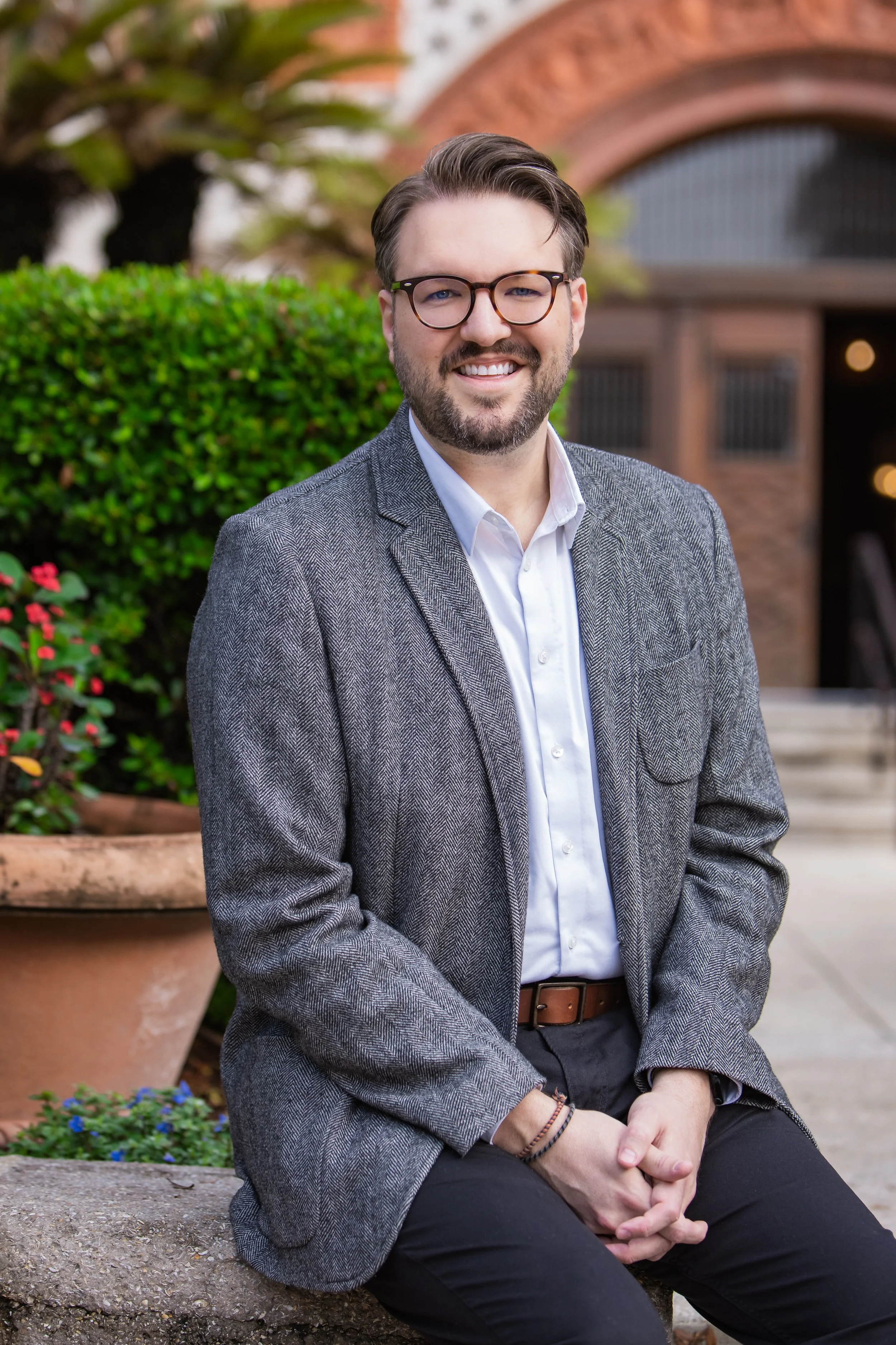 A smiling man with glasses, wearing a gray blazer and white shirt, sitting on a stone ledge outside a brick building with greenery and flowers in the background.