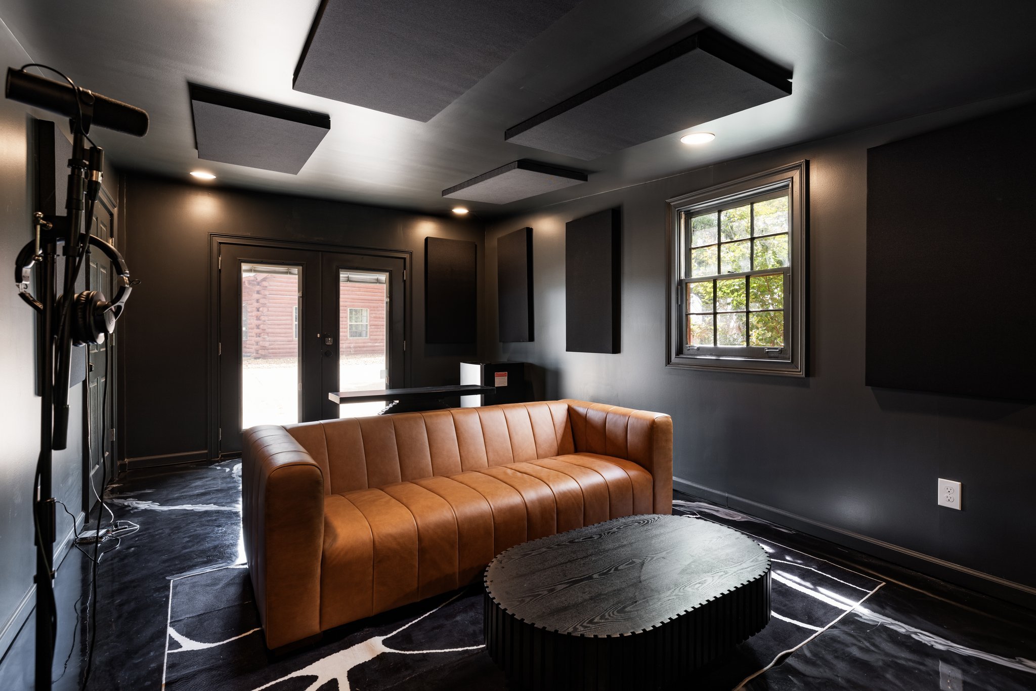 A recording studio with dark walls and ceiling, brown leather sofa, black patterned rug, window with green trees outside, and professional audio equipment including headphones and a microphone.