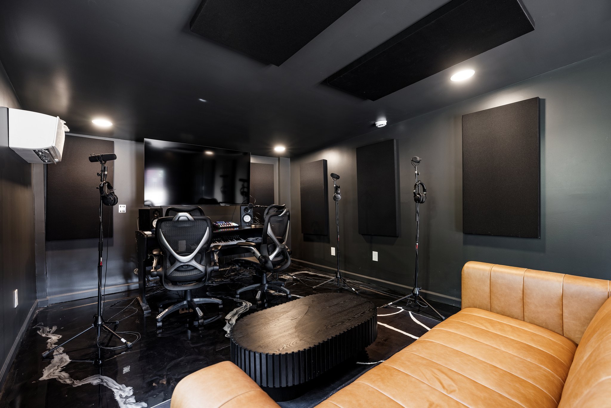 Recording studio room with black walls, acoustic panels, a large flat-screen TV, two office chairs, a mixing console, several microphone stands, a leather sofa, and a coffee table.