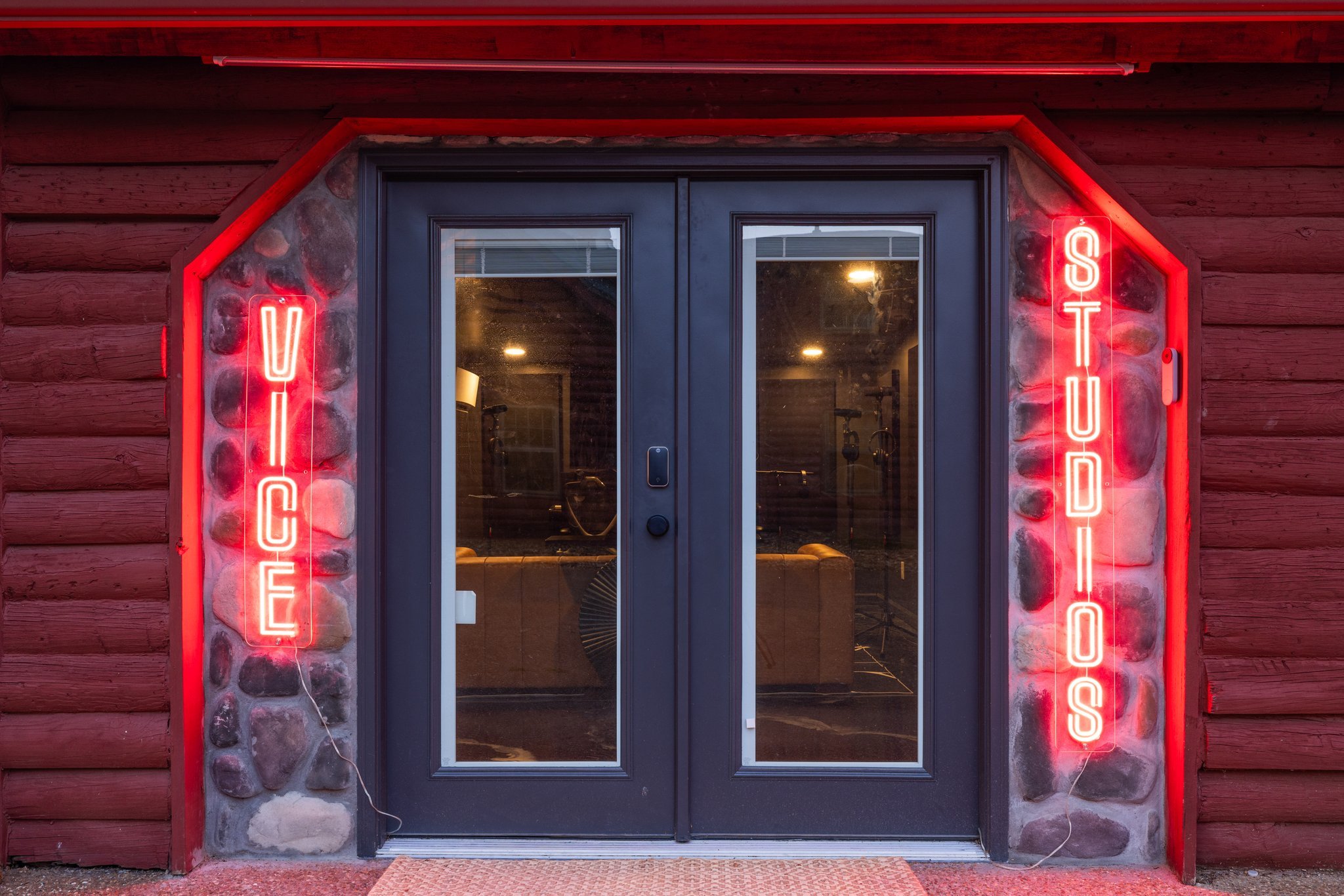 Glass doors of a yoga studio with neon red signs on either side that read 'YOGA' and 'STUDIOS.' The entrance is framed by a stone and wood exterior, illuminated by interior lighting.