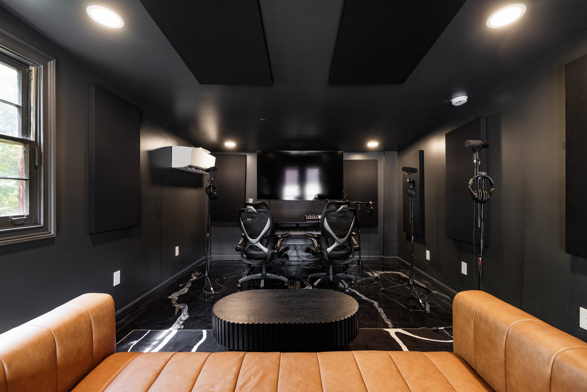 Modern recording studio with a large television screen, two computer chairs, microphones on stands, acoustic panels, and a brown leather couch.
