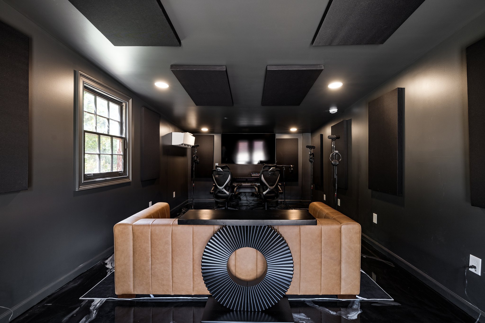 A soundproof music or recording studio with black walls, acoustic panels on the ceiling and walls, a window on the left, a large television or monitor on the back wall, two chairs at a mixing console, three microphone stands, and a tan sofa in the fo