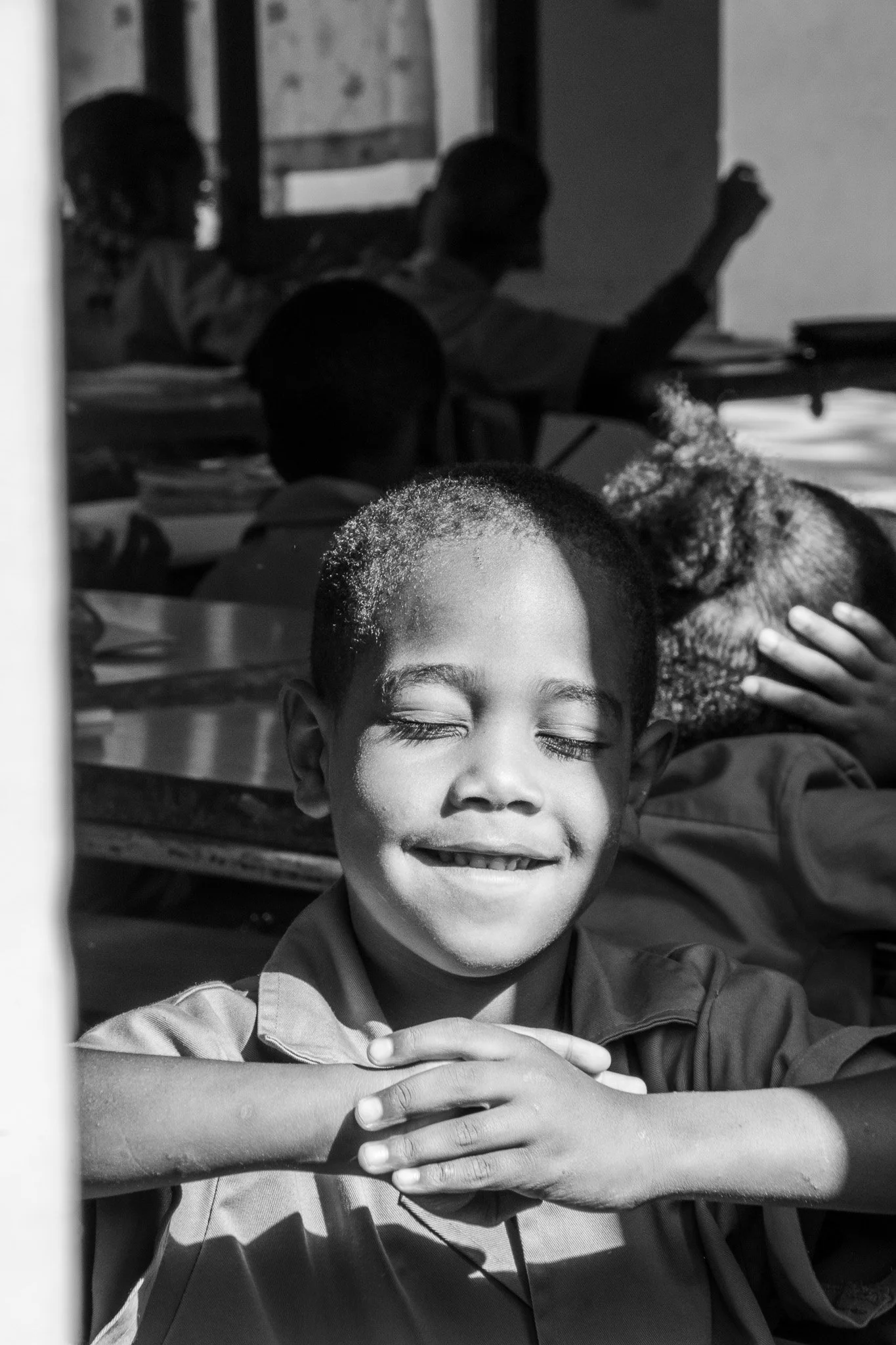 Ein glückliches Kind mit verschlossenen Augen, das in einem Klassenzimmer sitzt, während andere Kinder im Hintergrund lesen und Lernen.