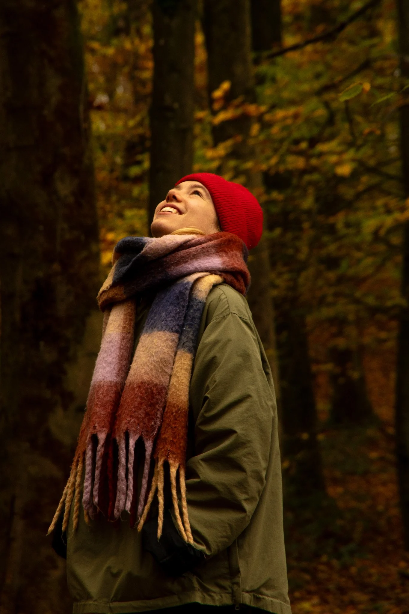 Eine Frau in einem Park im Herbst, trägt einen roten Hut und einen bunten Schal, schaut nach oben und scheint glücklich zu sein.