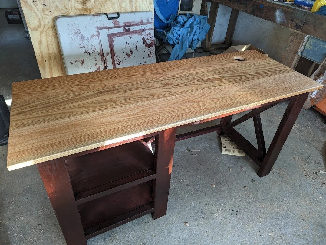 Wooden desk with a small hole in the corner, set in a workshop with tools and boards in the background.