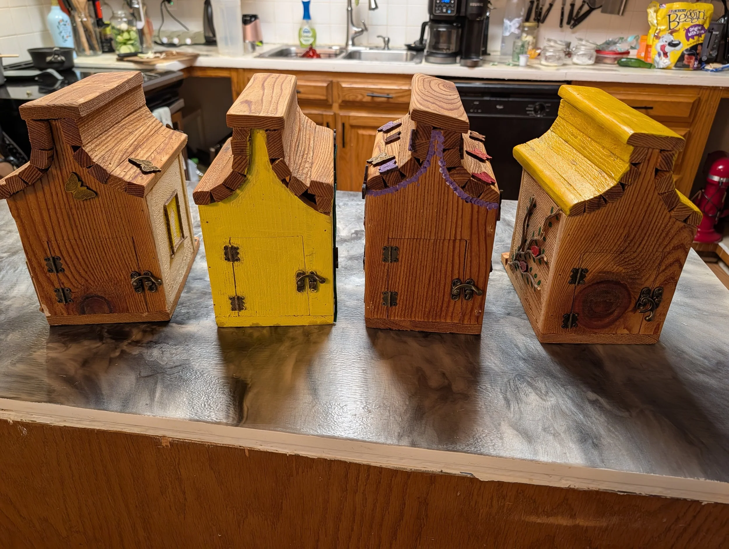 Four decorative wooden birdhouses with painted roofs and small decorations on a kitchen countertop.