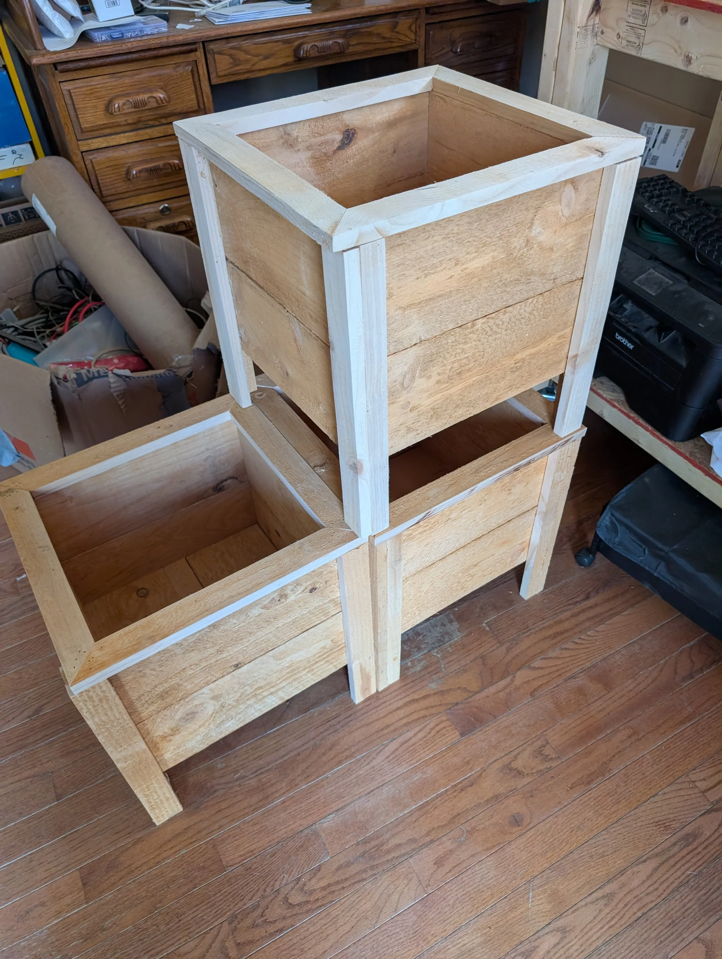 Unfinished wooden square planter box sitting on a workbench in a workshop. The box has a visible frame and interior, with some tools and wood pieces surrounding it.